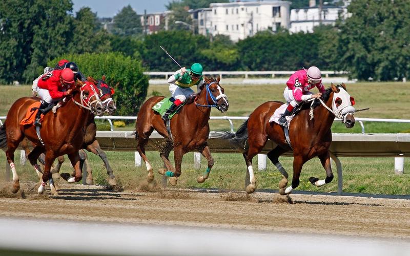 Três jóqueis disputam corrida de cavalos lado a lado em pista de turfe, com prédios ao fundo