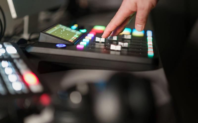 Mão de um operador ajustando os controles luminosos de uma mesa de corte (switcher) de transmissão de TV, com botões coloridos e tela de monitoramento