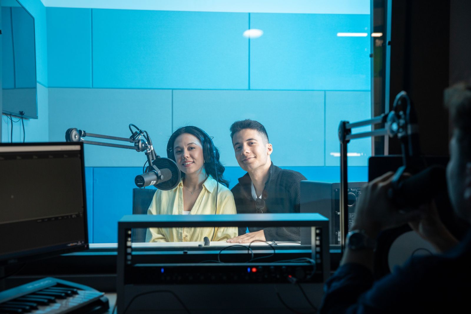 Dupla de apresentadores em estúdio de rádio moderno, usando fones de ouvido diante de um microfone profissional, com mesa de som e monitores ao fundo em ambiente iluminado de azul.
