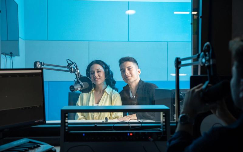 Dupla de apresentadores em estúdio de rádio moderno, usando fones de ouvido diante de um microfone profissional, com mesa de som e monitores ao fundo em ambiente iluminado de azul.