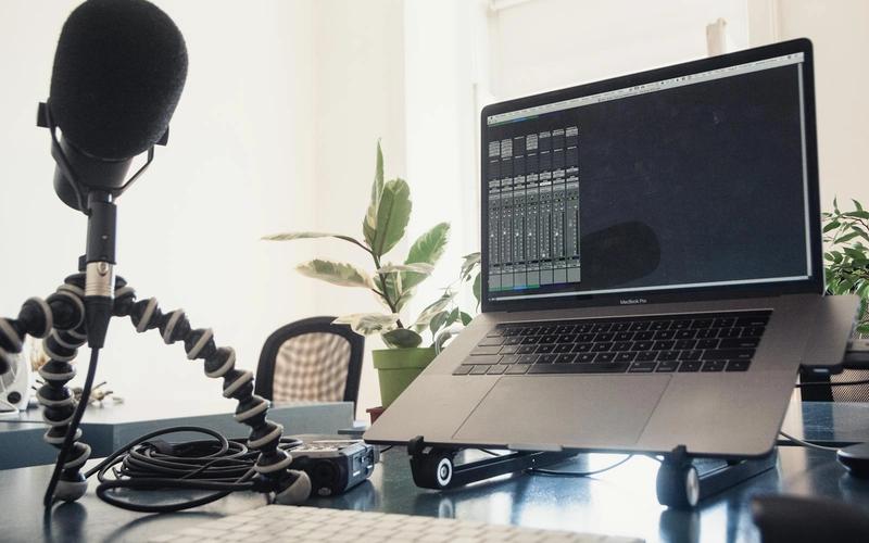 Setup de podcast em casa: microfone condensador em tripé flexível ao lado de um notebook com software de áudio na tela, sobre uma mesa com plantas e luz natural de janela.