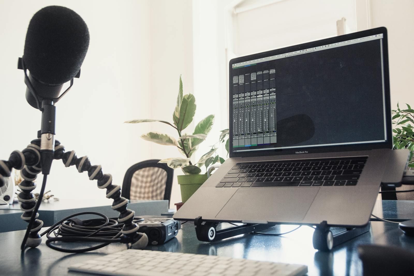 Setup de podcast em casa: microfone condensador em tripé flexível ao lado de um notebook com software de áudio na tela, sobre uma mesa com plantas e luz natural de janela.