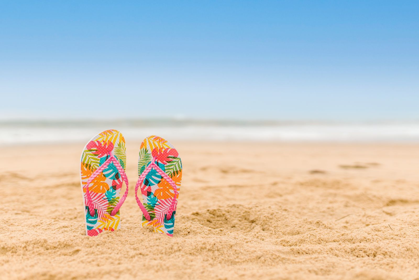 Par de chinelos de dedo com estampa tropical colorida (rosa, laranja, verde e amarelo) fincado na areia de uma praia, com céu azul limpo ao fundo.