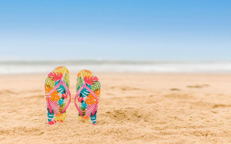 Par de chinelos de dedo com estampa tropical colorida (rosa, laranja, verde e amarelo) fincado na areia de uma praia, com céu azul limpo ao fundo.
