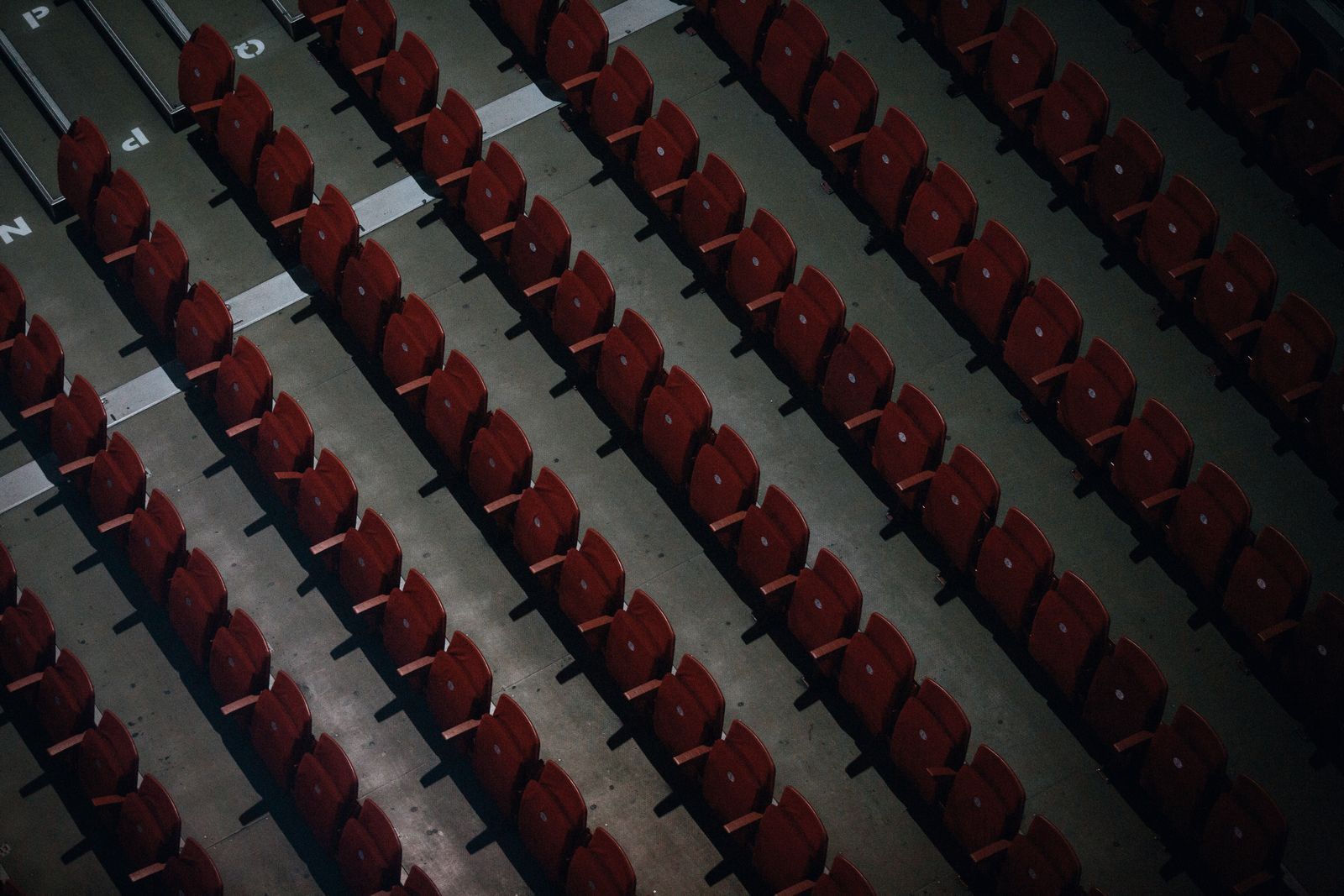 Vista aérea de fileiras de cadeiras vermelhas vazias em arquibancada de estádio, com luz e sombra formando um padrão diagonal