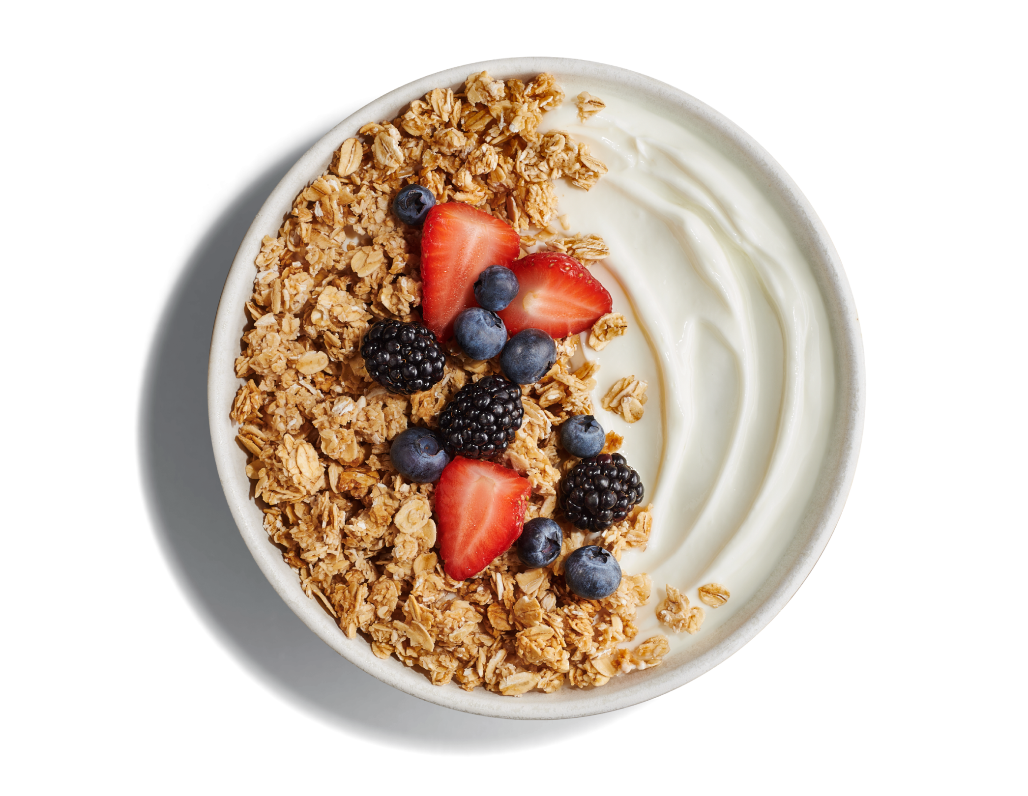 White bowl of plain yogurt with golden, crunchy granola topped with cut strawberries, blackberries and blueberries on a bright blue background.