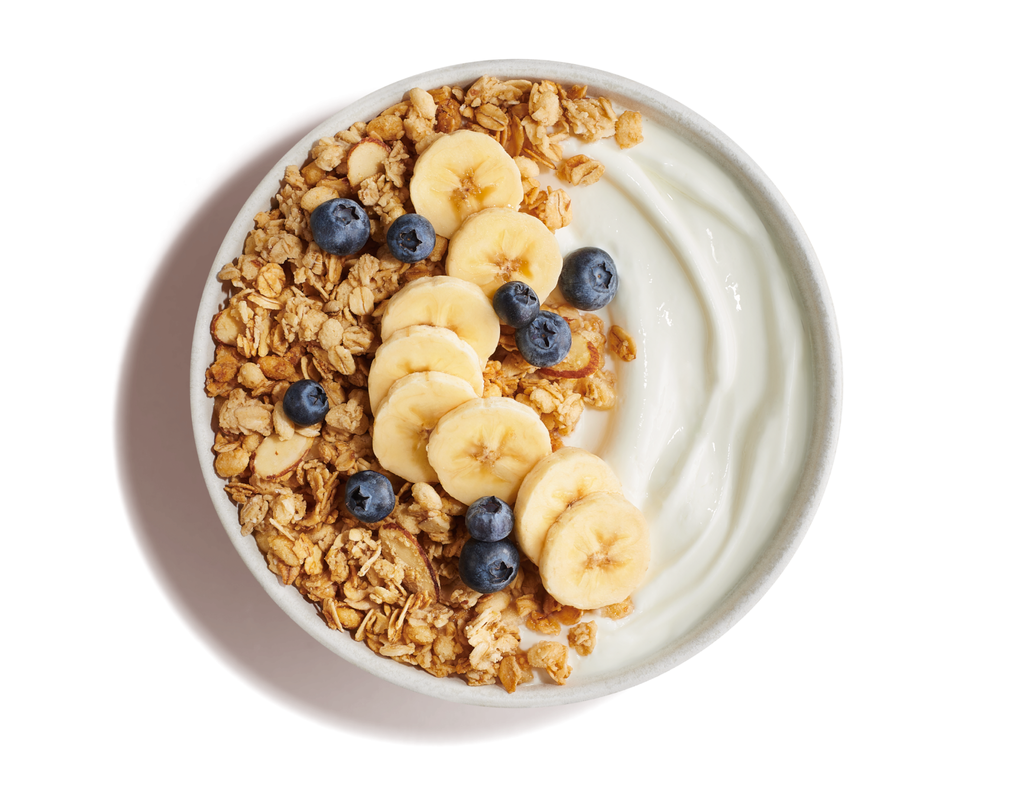 White bowl of plain yogurt with golden crunchy granola topped with banana slices and blueberries on a yellow background.