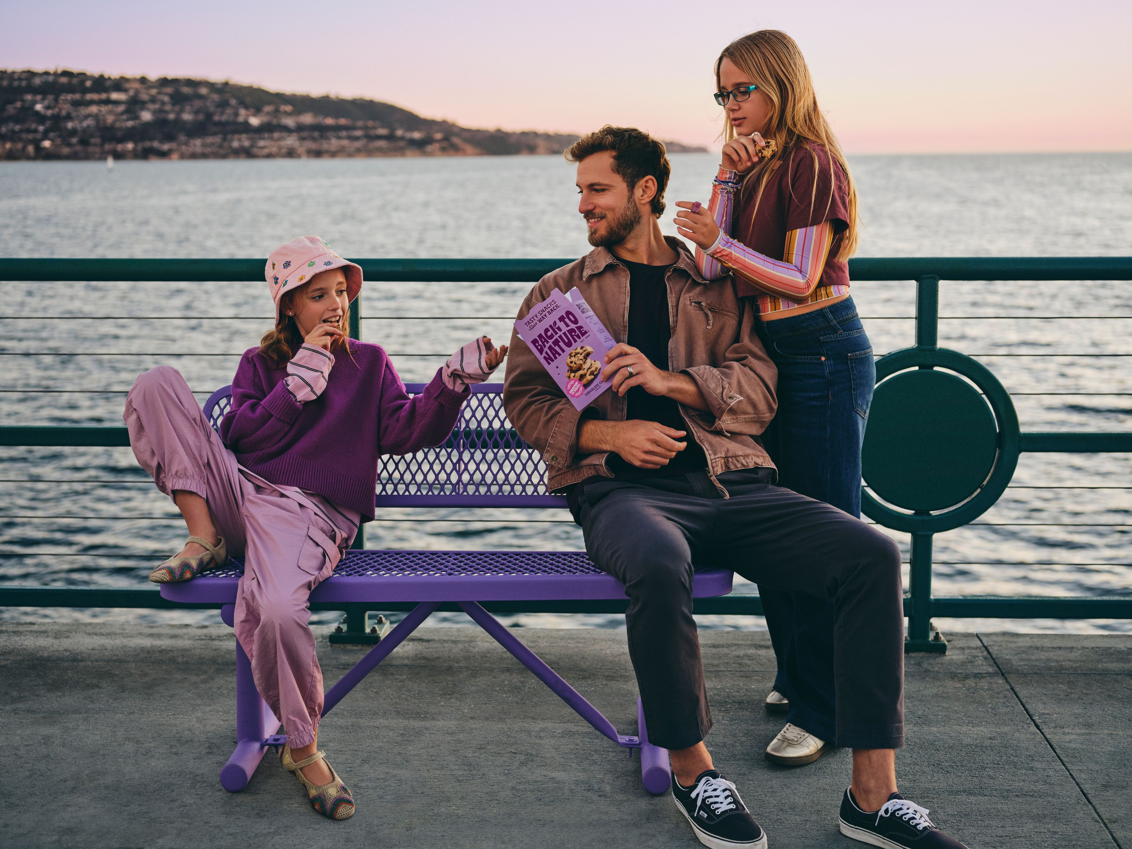 A father and 2 daughters sitting on a bench by the pier, eating cookies