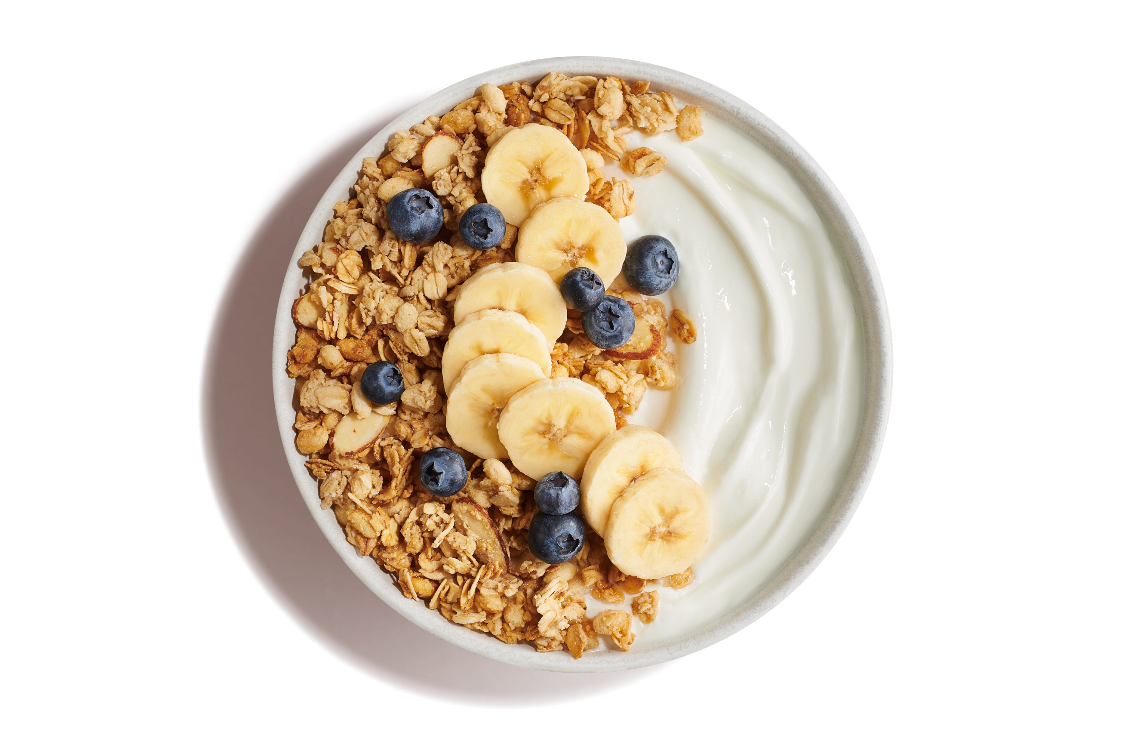 White bowl of plain yogurt with golden crunchy granola topped with banana slices and blueberries on a yellow background.