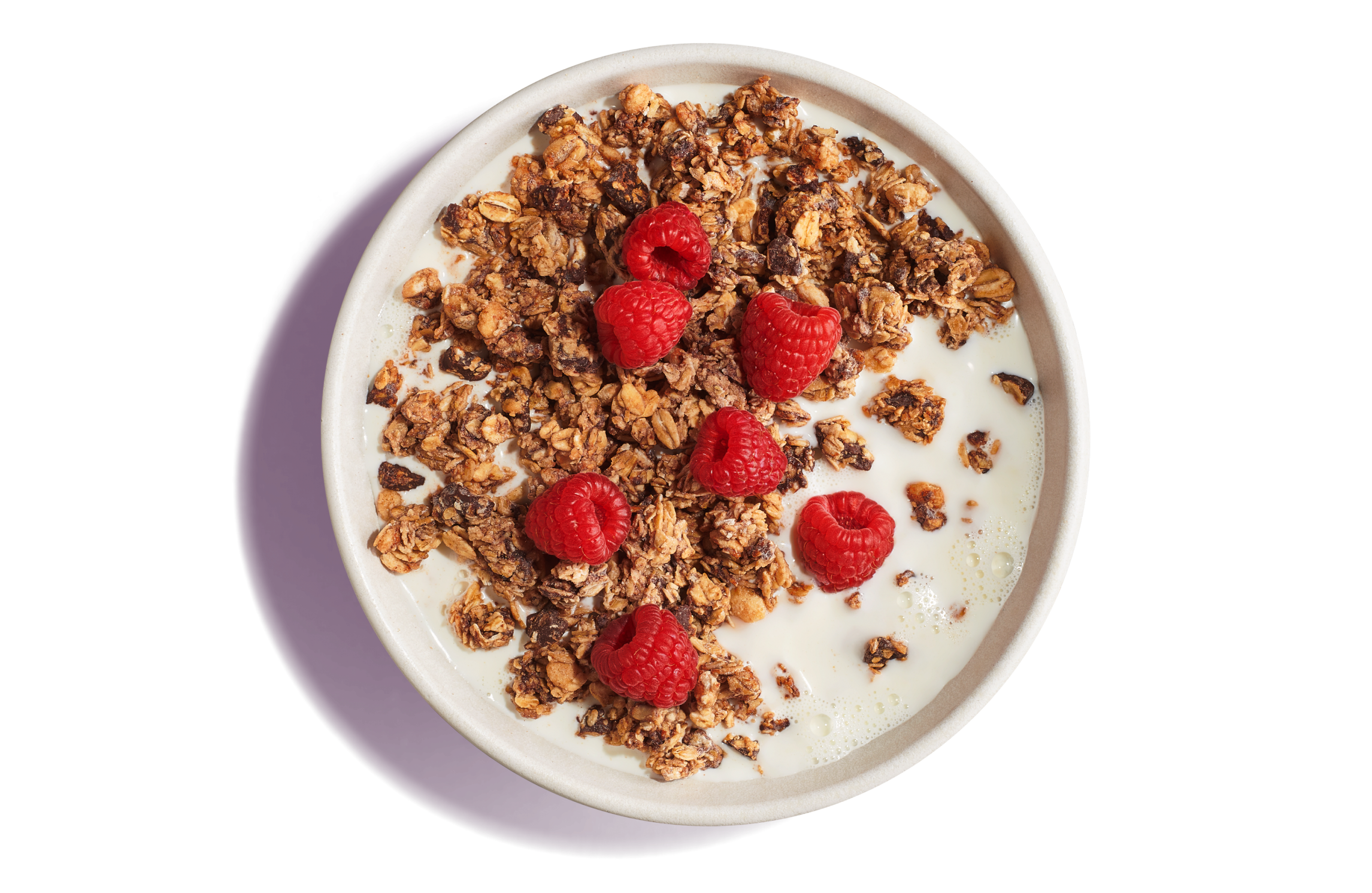 White bowl of milk with dark brown, crunchy granola topped with whole raspberries on a pruple background.  