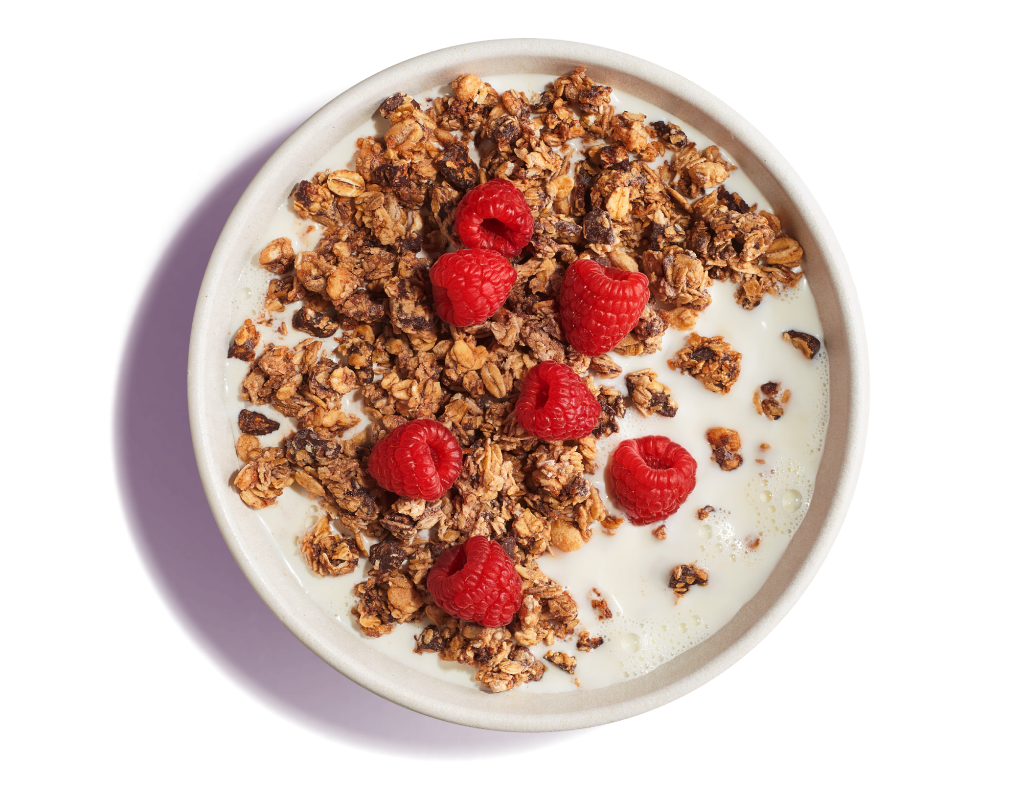 White bowl of milk with dark brown, crunchy granola topped with whole raspberries on a pruple background.