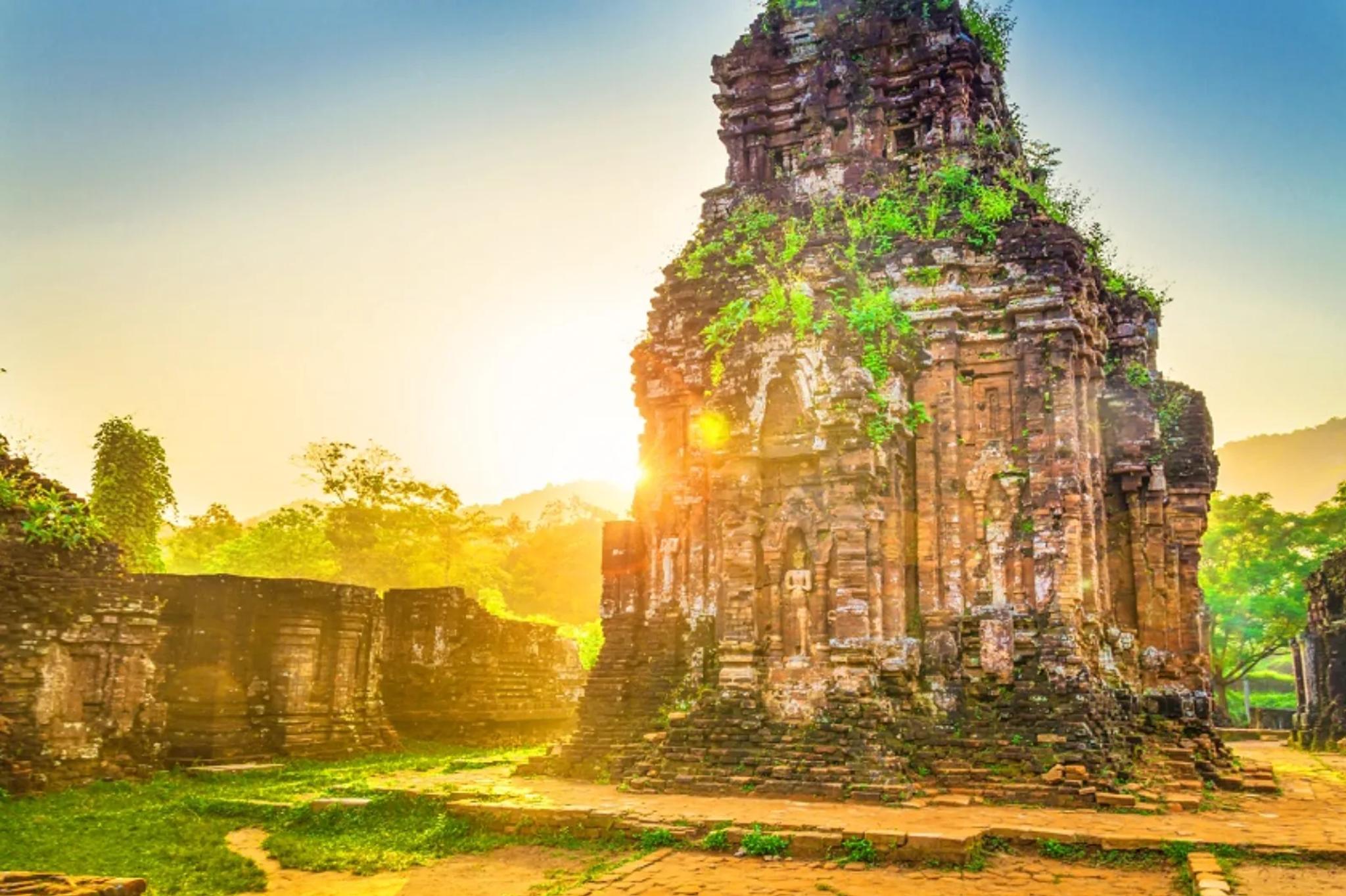 Sunrise light flaring over a Cham tower with grass growing on the ruins at My Son Sanctuary.