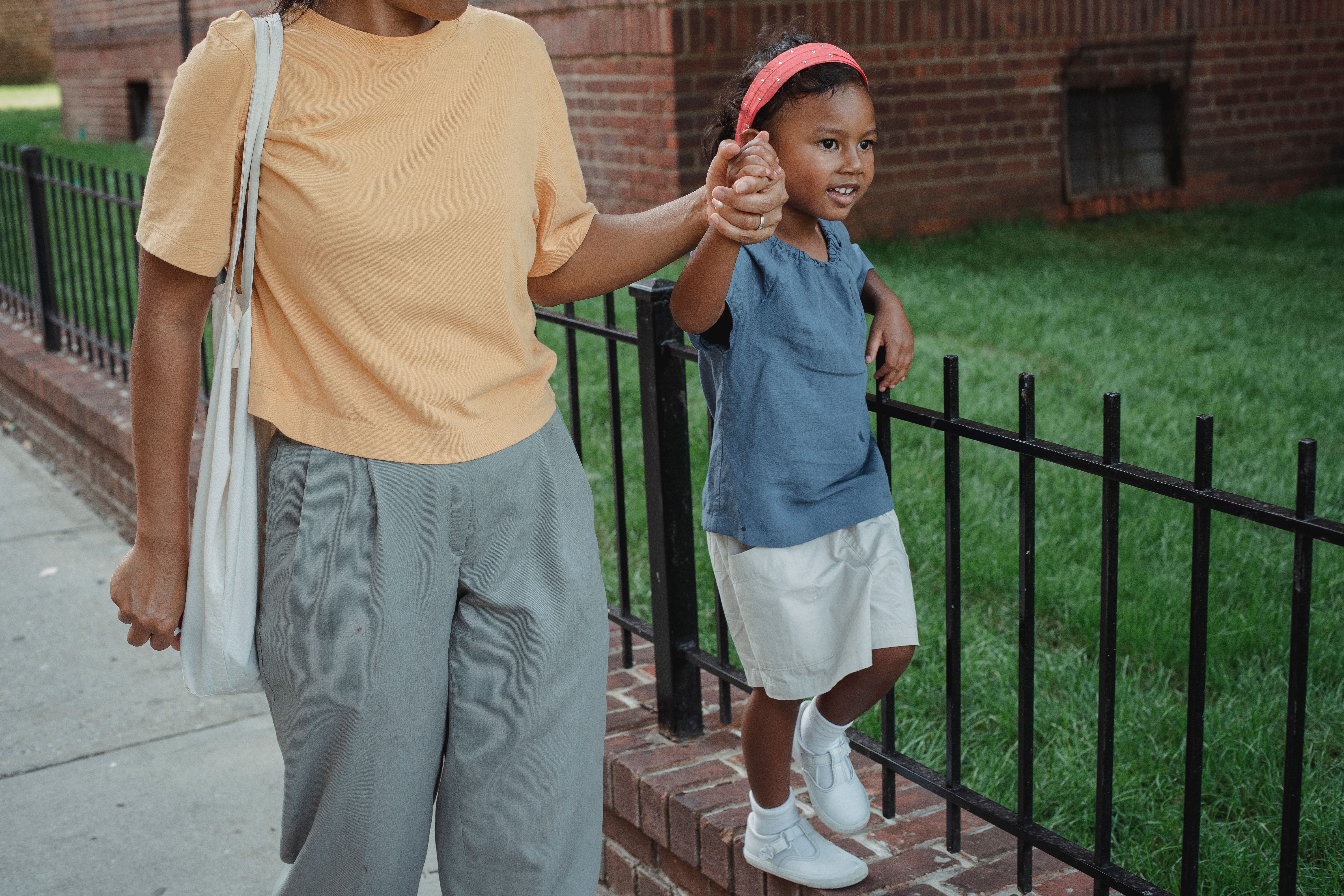 Image A mother holds her young daughter's hand as they walk together on a city sidewalk on the way to school.