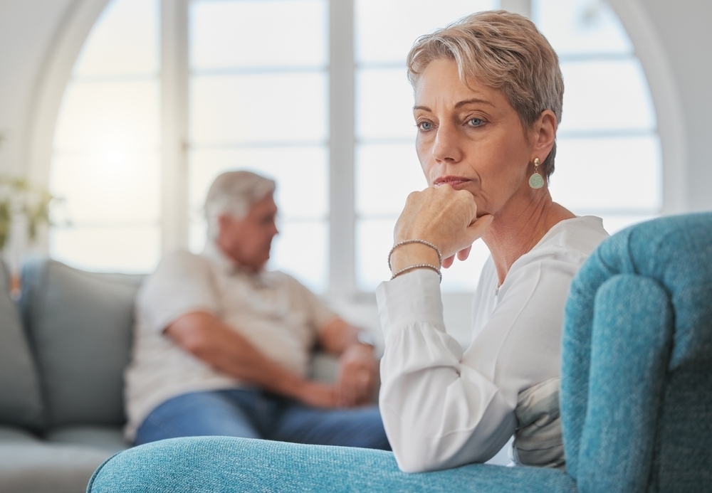 Image Woman in her 50s sitting thoughtfully in a living room with older husband blurred in background, representing grey divorce in Illinois.