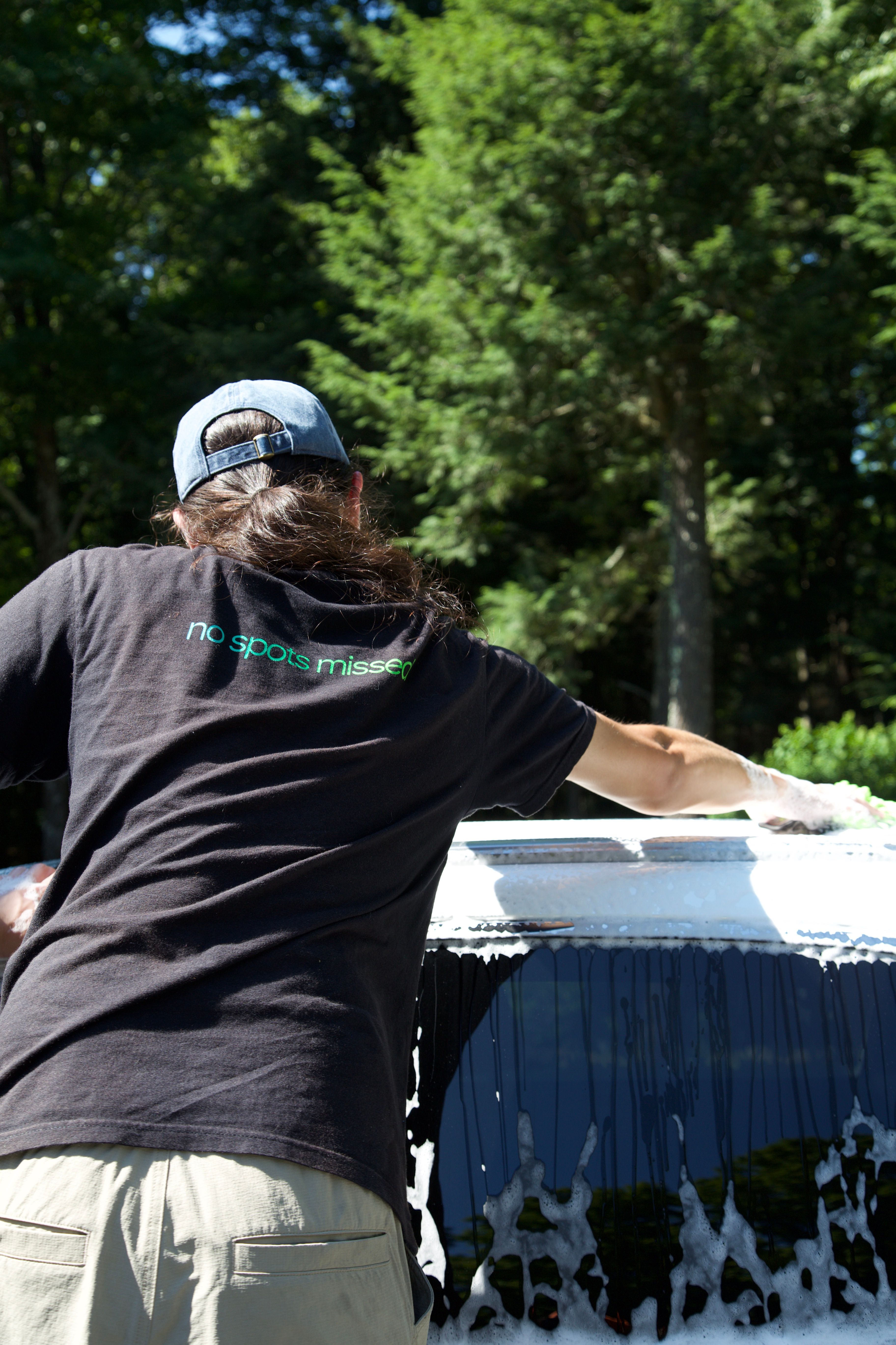 A person facing away from the camera using a wash mitt to scrub a car. The black shirt says "no spots missed".