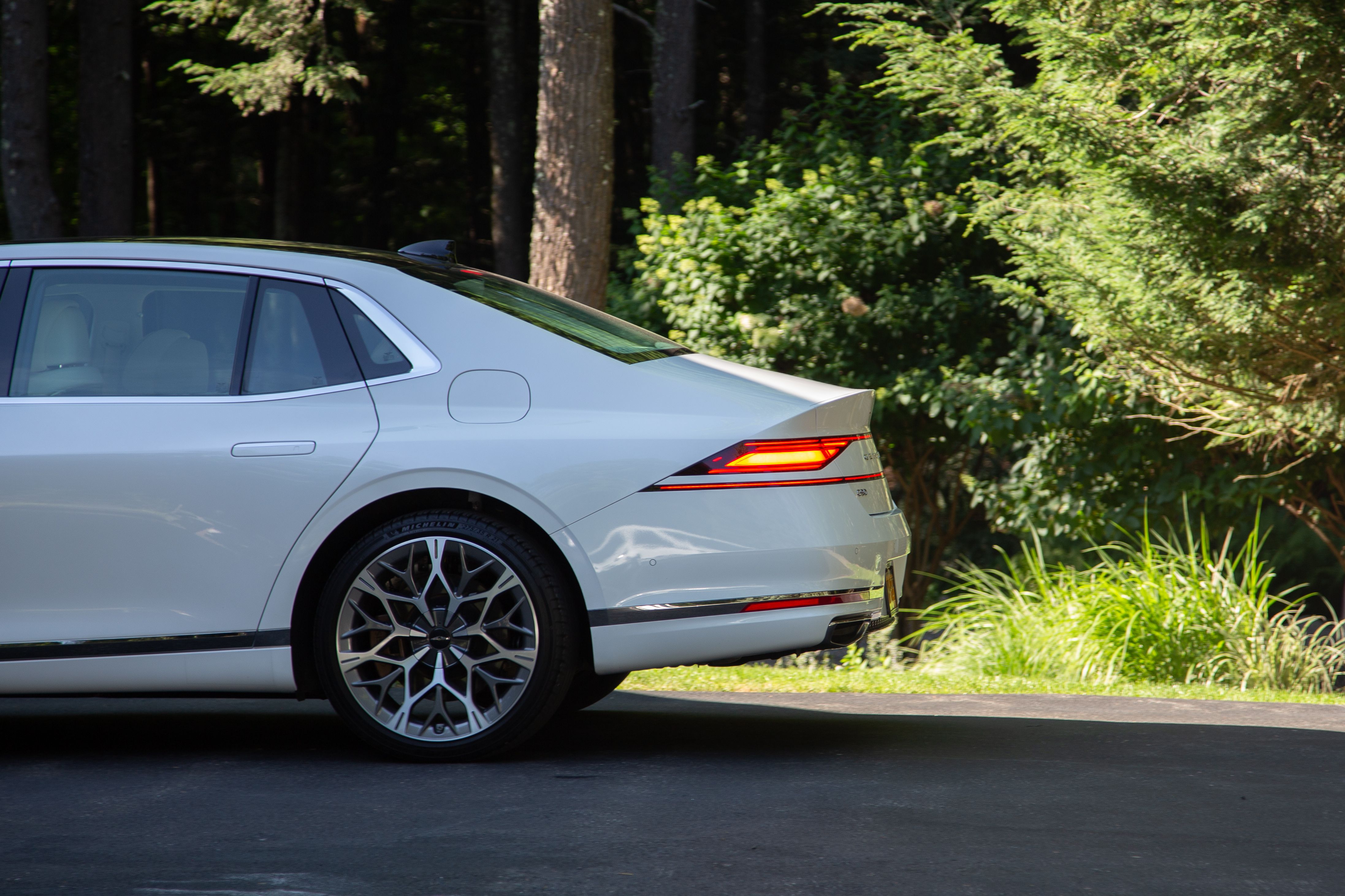 The back half of a ceramic coated Genesis G90 in a driveway with the tail lights glowing red. The reflections of green trees can be see in the paint.