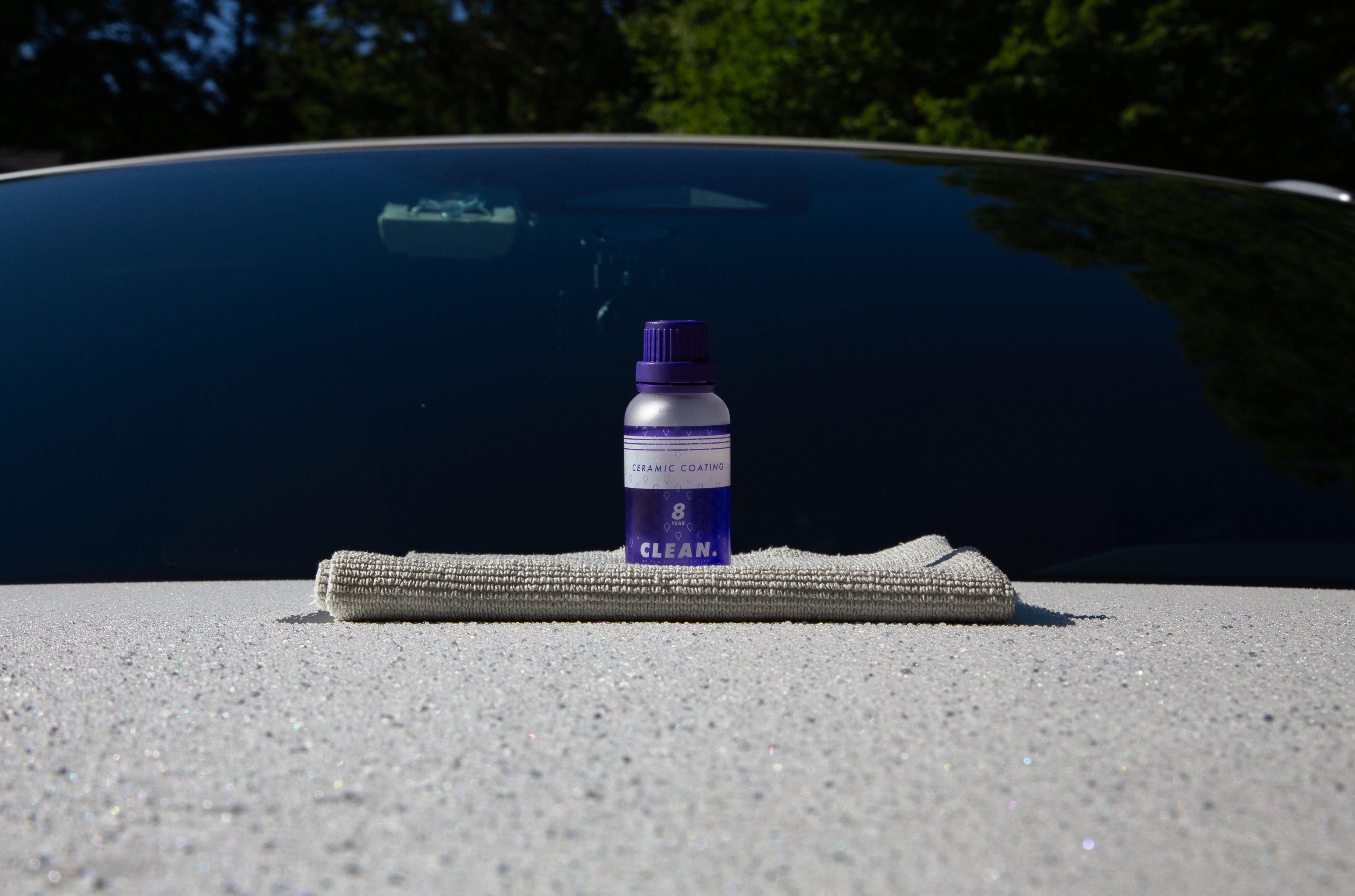 A Ceramic Coating bottle sitting on top of a gray microfiber cloth, neatly folded, on top of a white car hood. The car hood has small water droplet beads, formed from the ceramic coating.