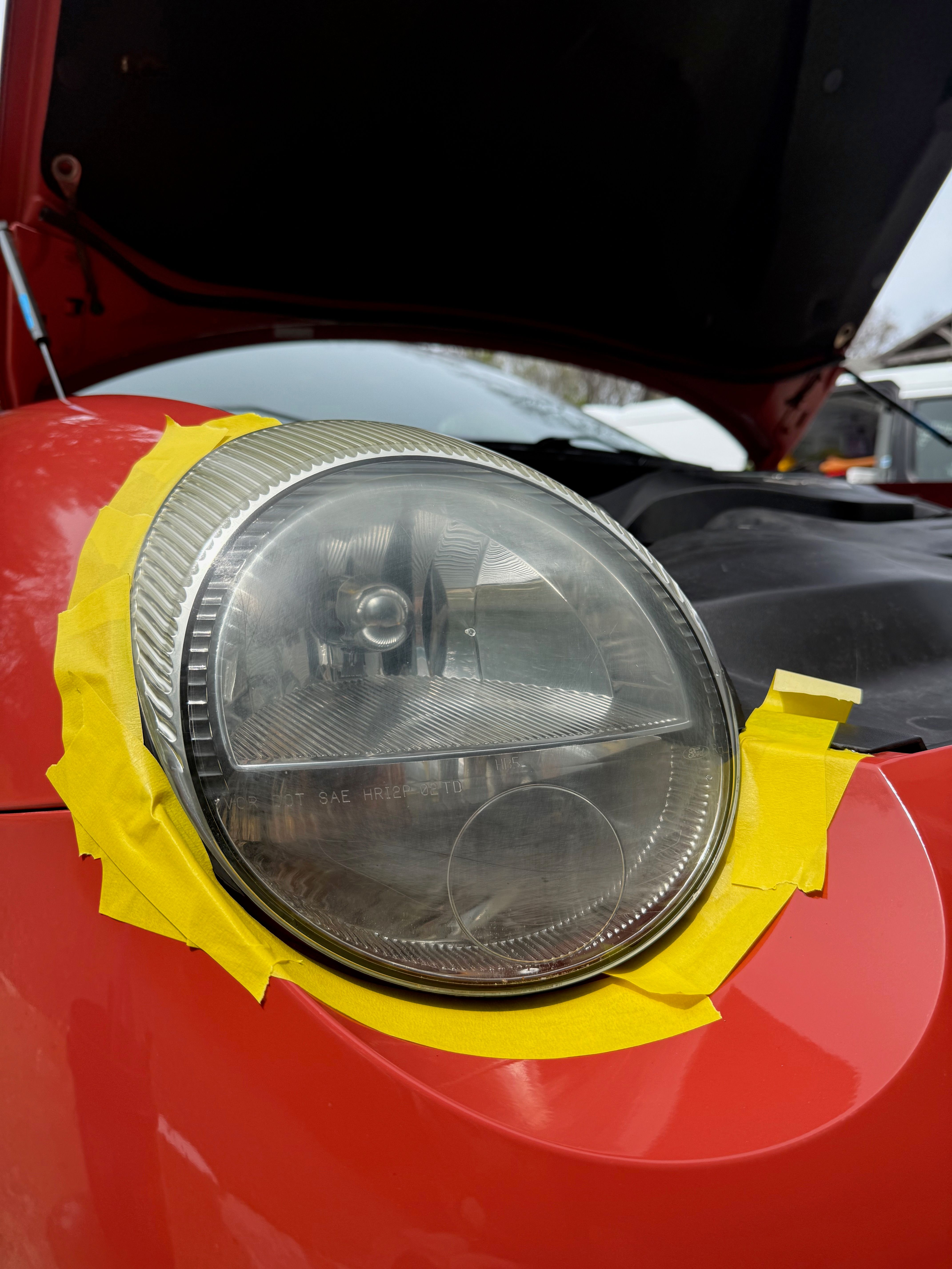 A cloudy, oxidized headlight on a Ford Thunderbird with orange paint. The hood is open and the headlight is in focus, with yellow tape surrounding it to mask the area.
