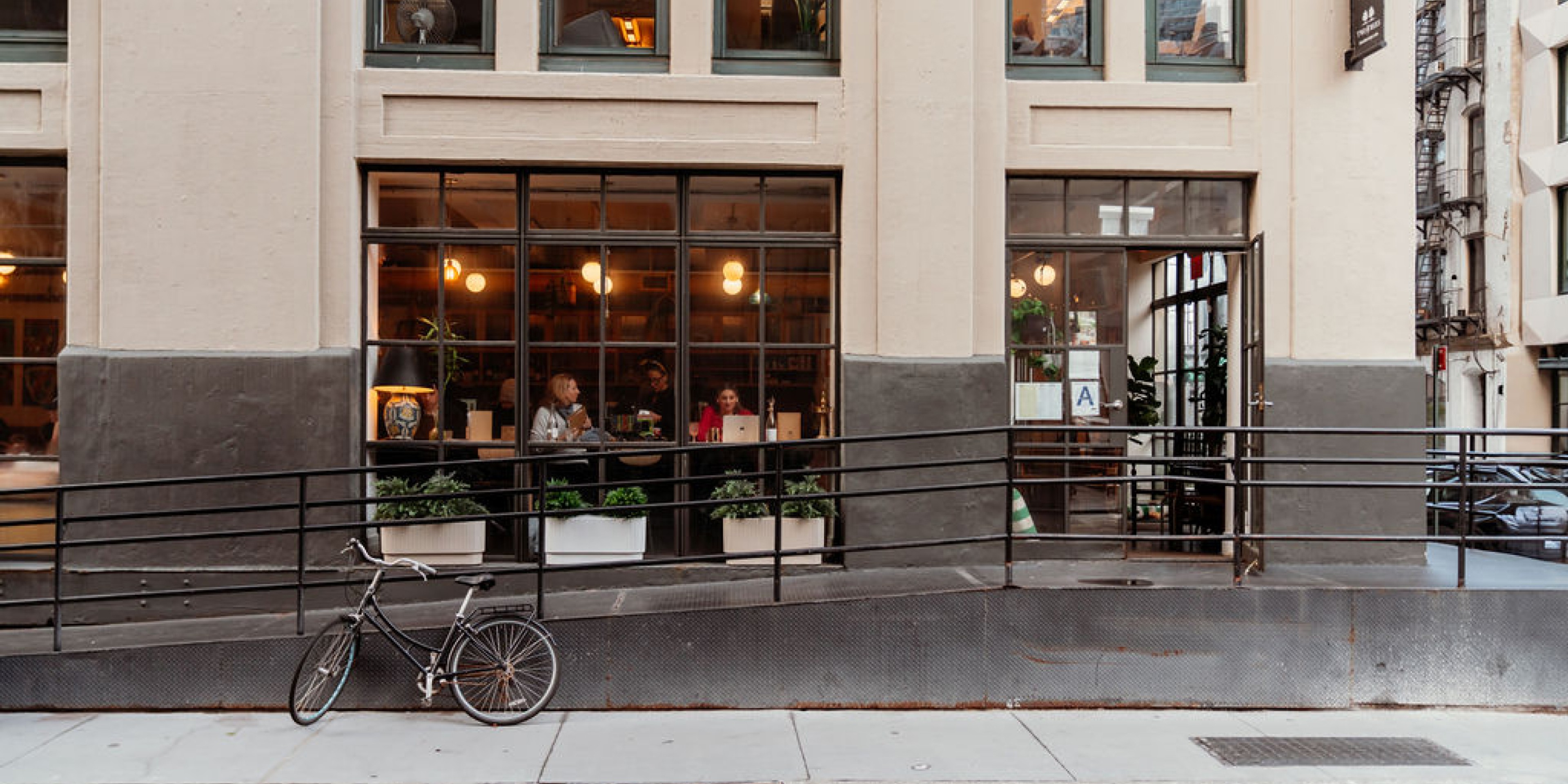 Urban café exterior with large windows, warm interior lighting, visible diners, potted plants, and a bicycle parked along the sidewalk in a city setting.