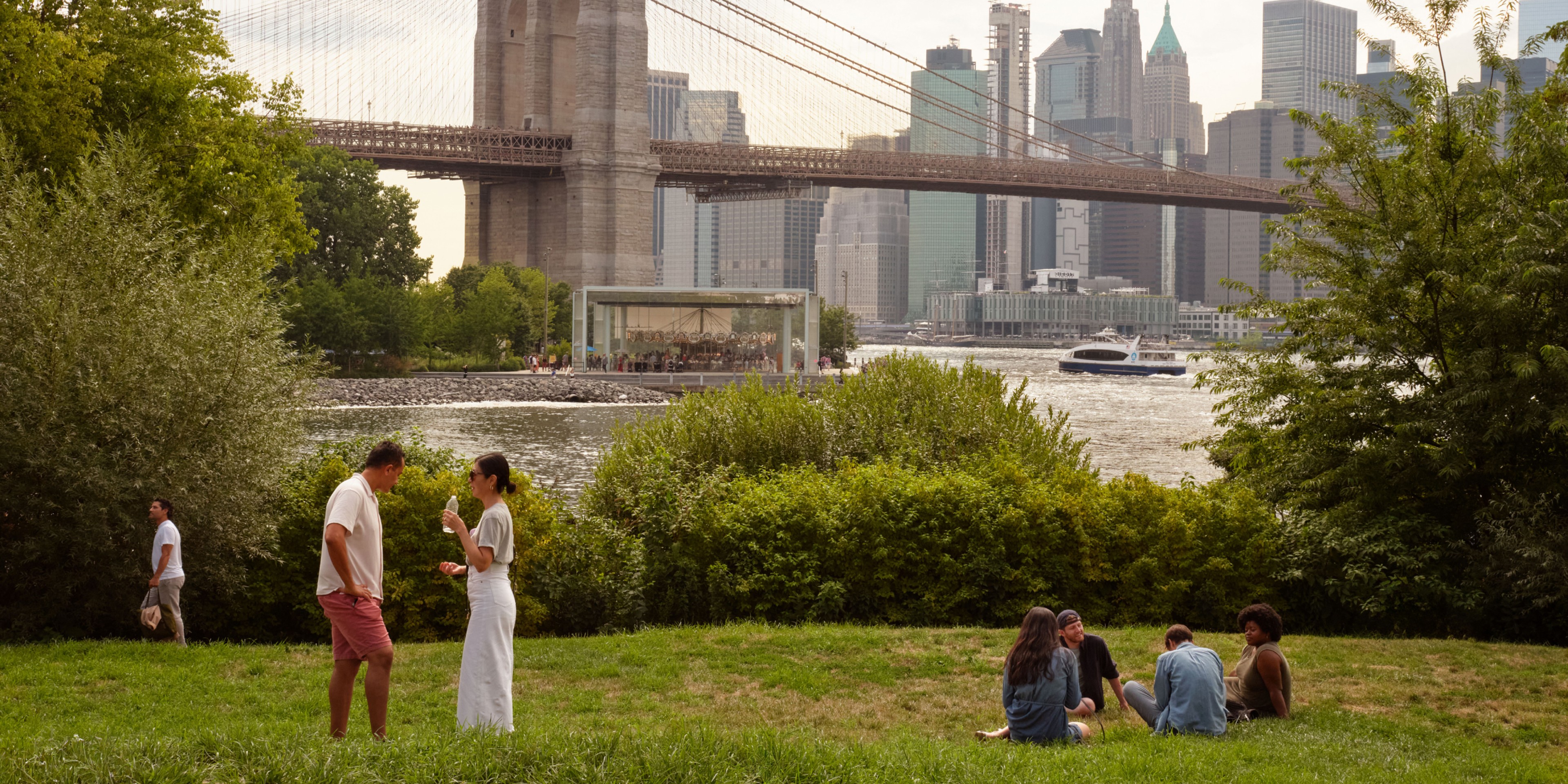 Groups of people enjoying a sunny day at a waterfront park with the Brooklyn Bridge, Jane’s Carousel, and downtown Manhattan skyline in the background.