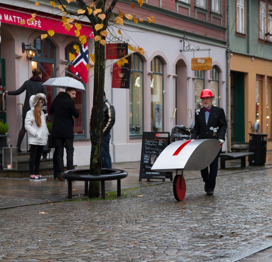 Bildet viser en mann som triller en slags trillebår og publikum som snur seg rundt for å se etter ham.