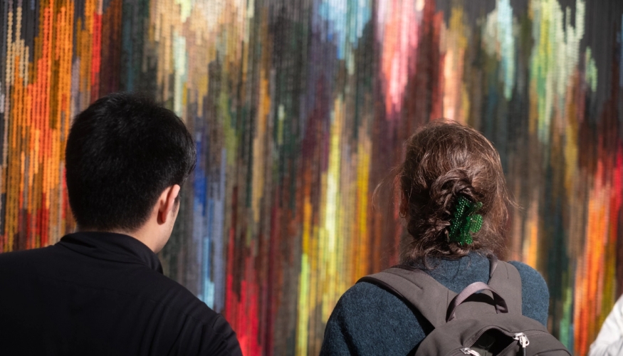 To mennesker står foran et stort tekstilverk i mange farger