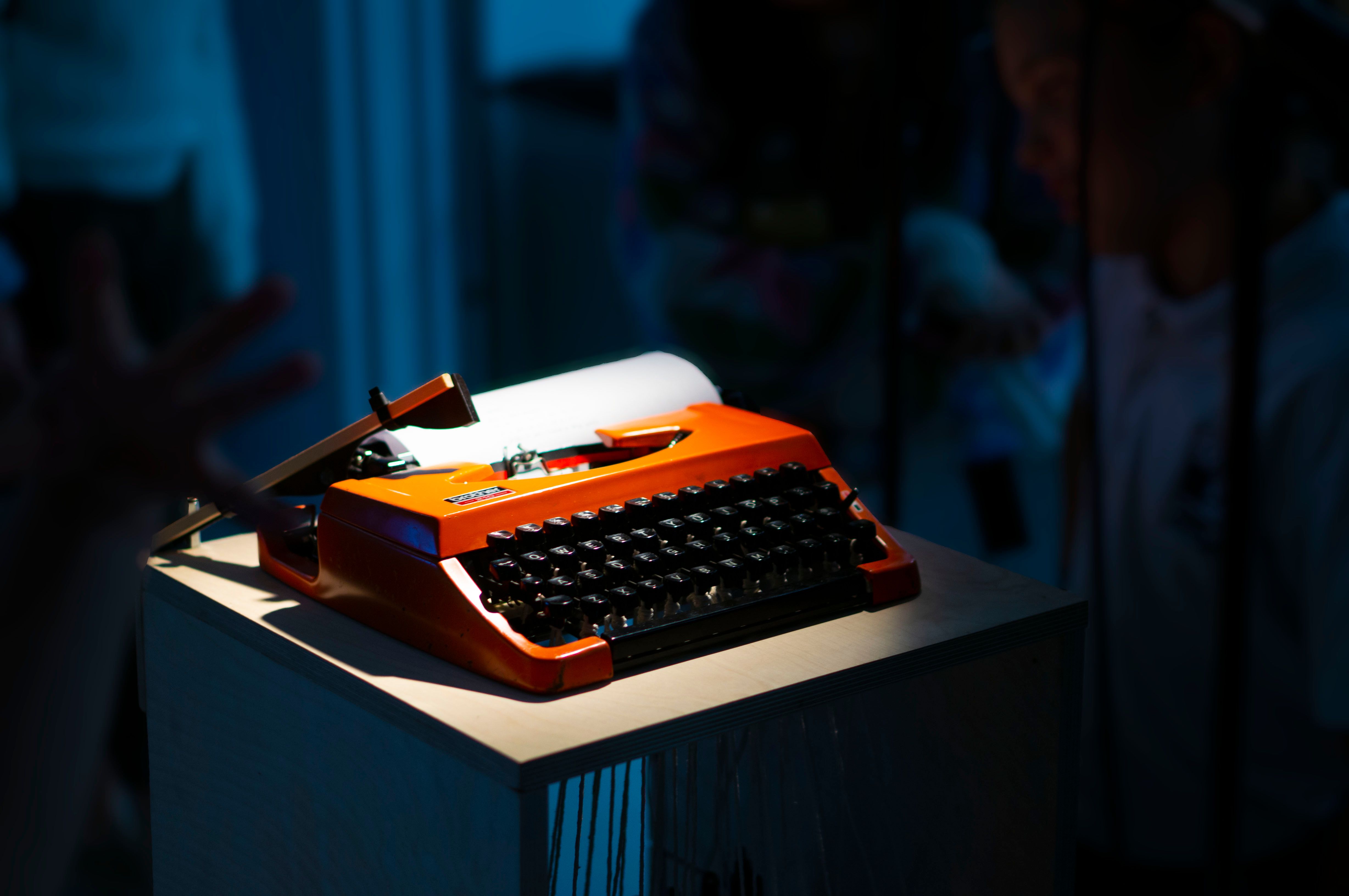 dramaticaly lit orange typewriter, with people looking at it in the background