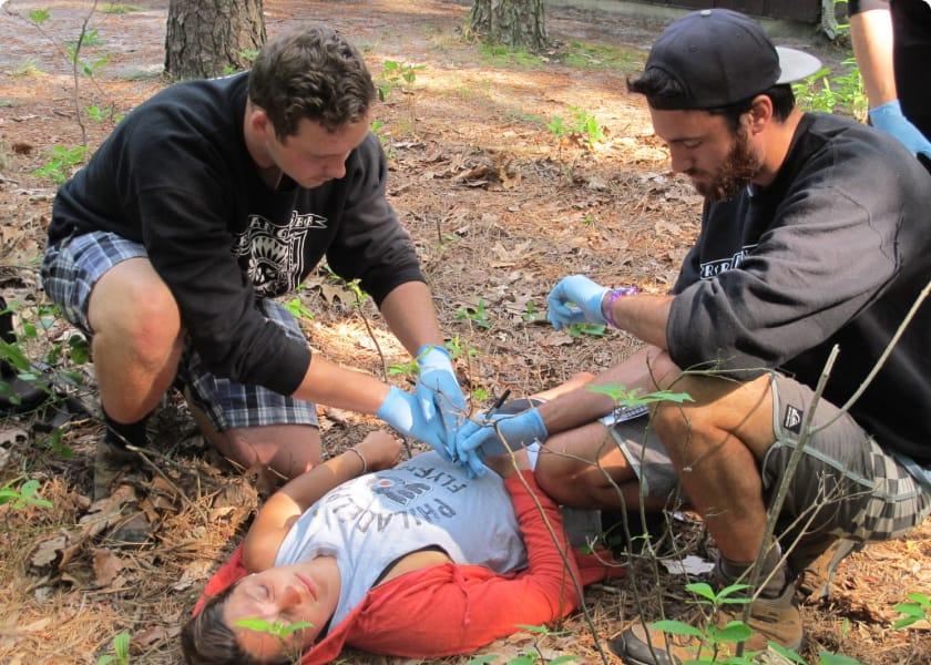 Two men with surgical gloves in forest applying medical attention to woman laying on ground.