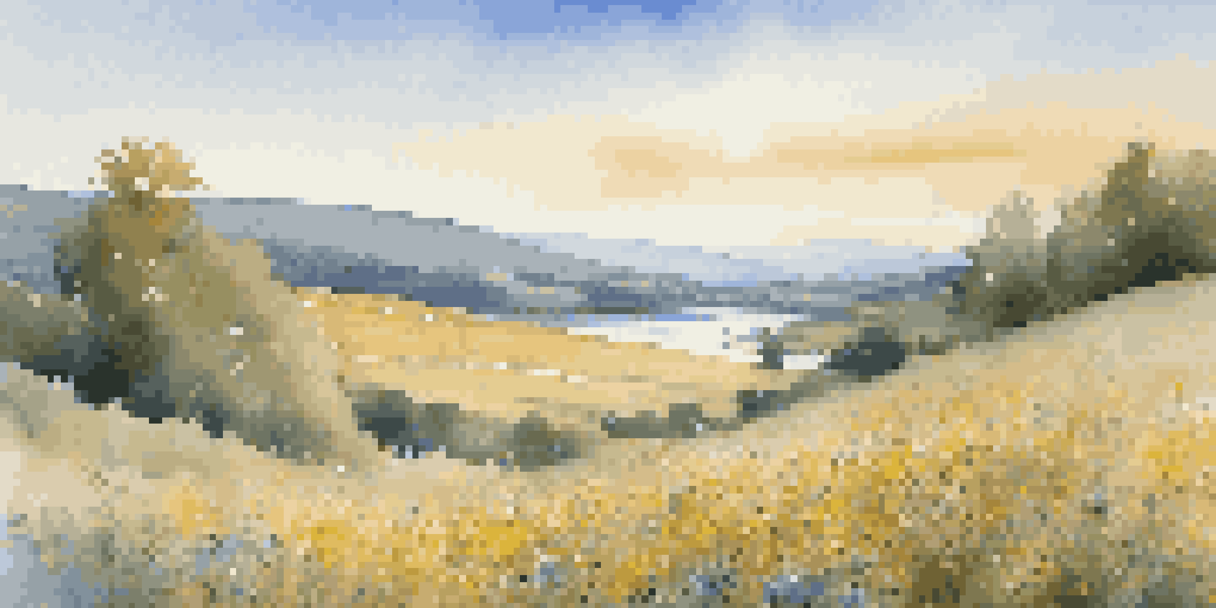 A colorful field of wildflowers under a bright blue sky with a calm river flowing in the foreground and rolling hills in the background.