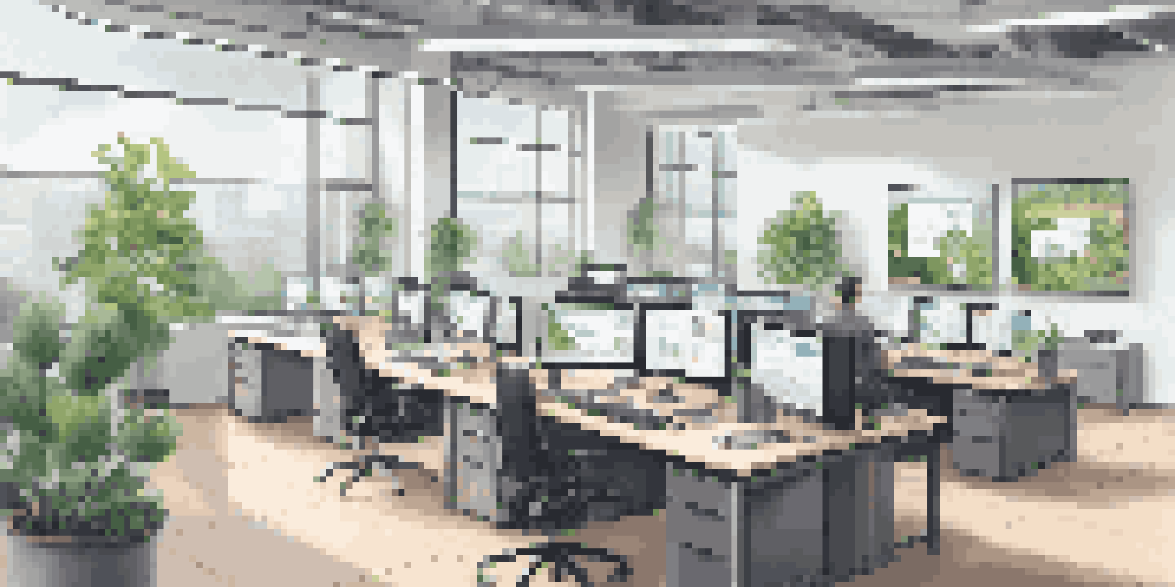 A well-lit modern office with multiple computer screens displaying digital management software, plants on desks, and natural light coming through large windows.