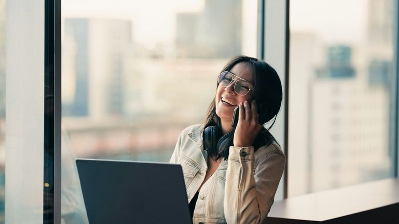 A woman on a phone call near a window with a laptop beside her