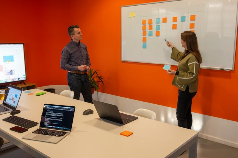 Two people discussing work in front of a whiteboard covered in sticky notes