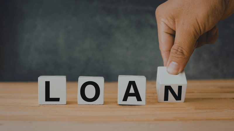 A person's hand placing the final letter block to complete the word loan on a wooden surface, illustrating the steps to apply for a small business loan.