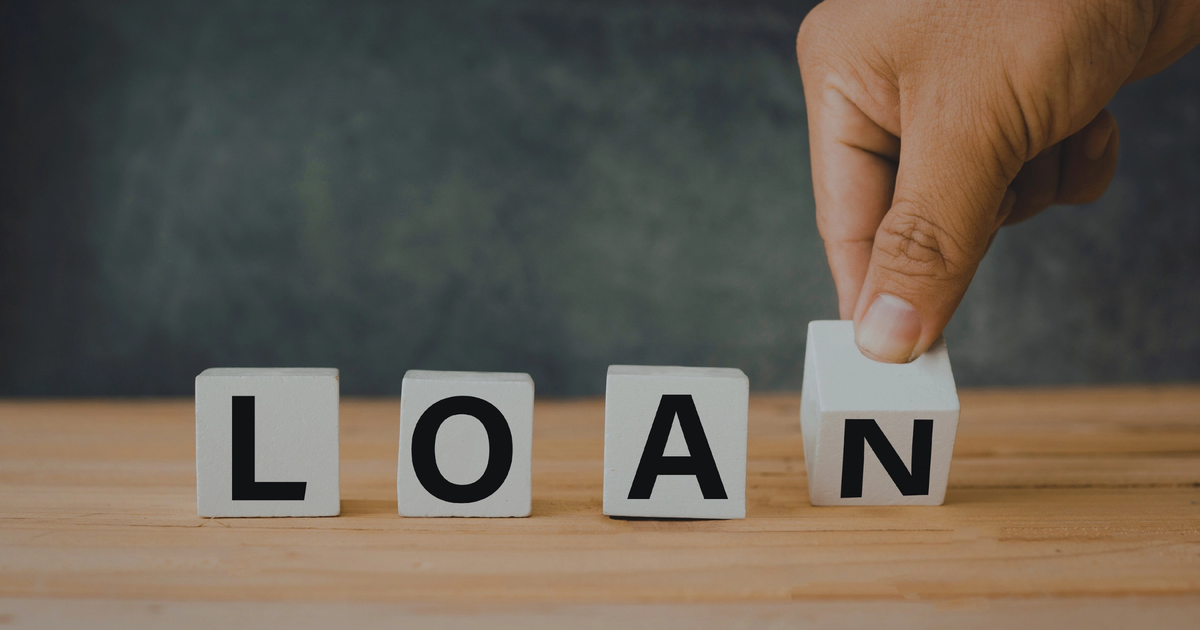 A person's hand placing the final letter block to complete the word loan on a wooden surface, illustrating the steps to apply for a small business loan.