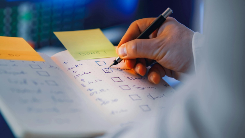 Lender writing a checklist for a commercial loan in a notebook with sticky notes visible in the background.
