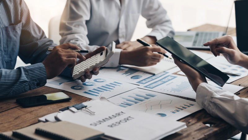 Two business professionals sitting at a desk reviewing financial documents, spreadsheets, and a calculator during a business meeting.