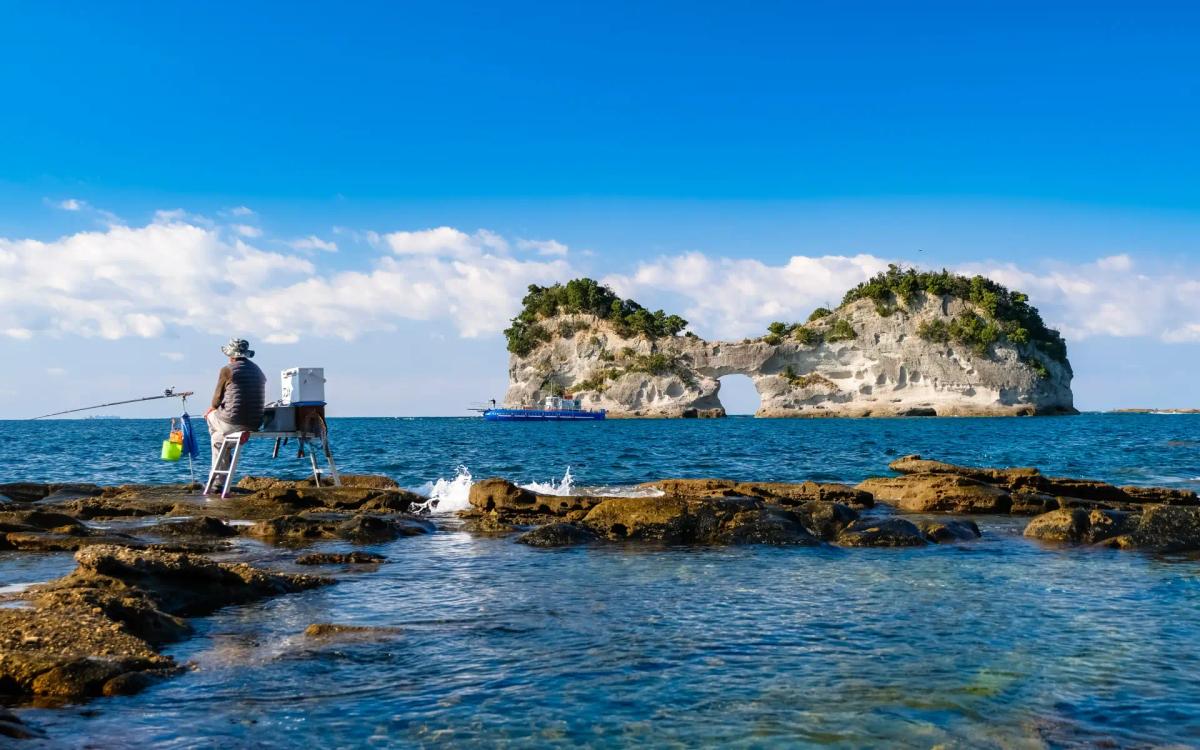 Fisherman sitting on rocks casting line toward island with blue ocean and clear sky