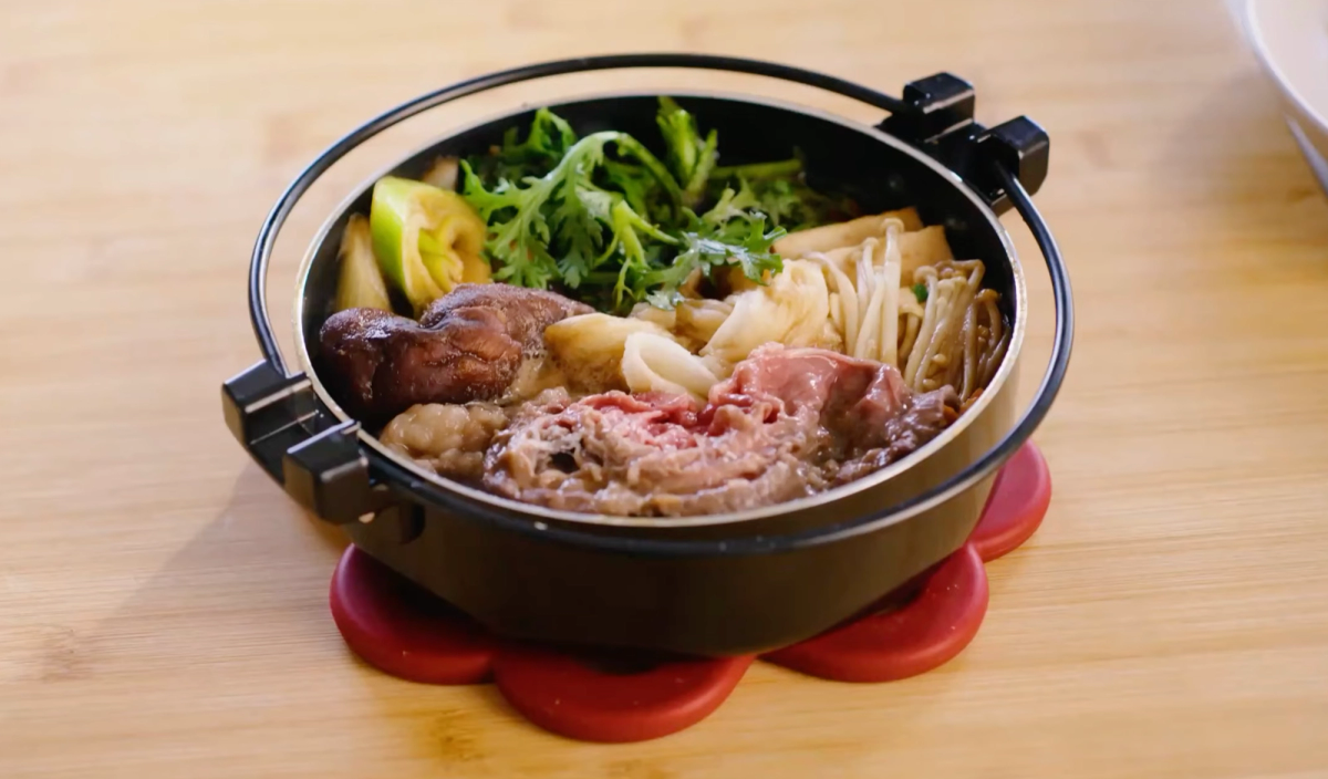 Hot pot with broth, beef, noodles, vegetables, and mushrooms on wooden table