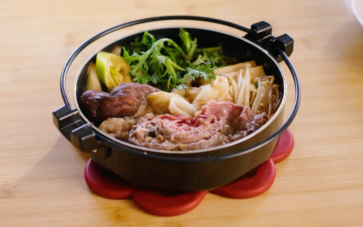 Hot pot with broth, beef, noodles, vegetables, and mushrooms on wooden table