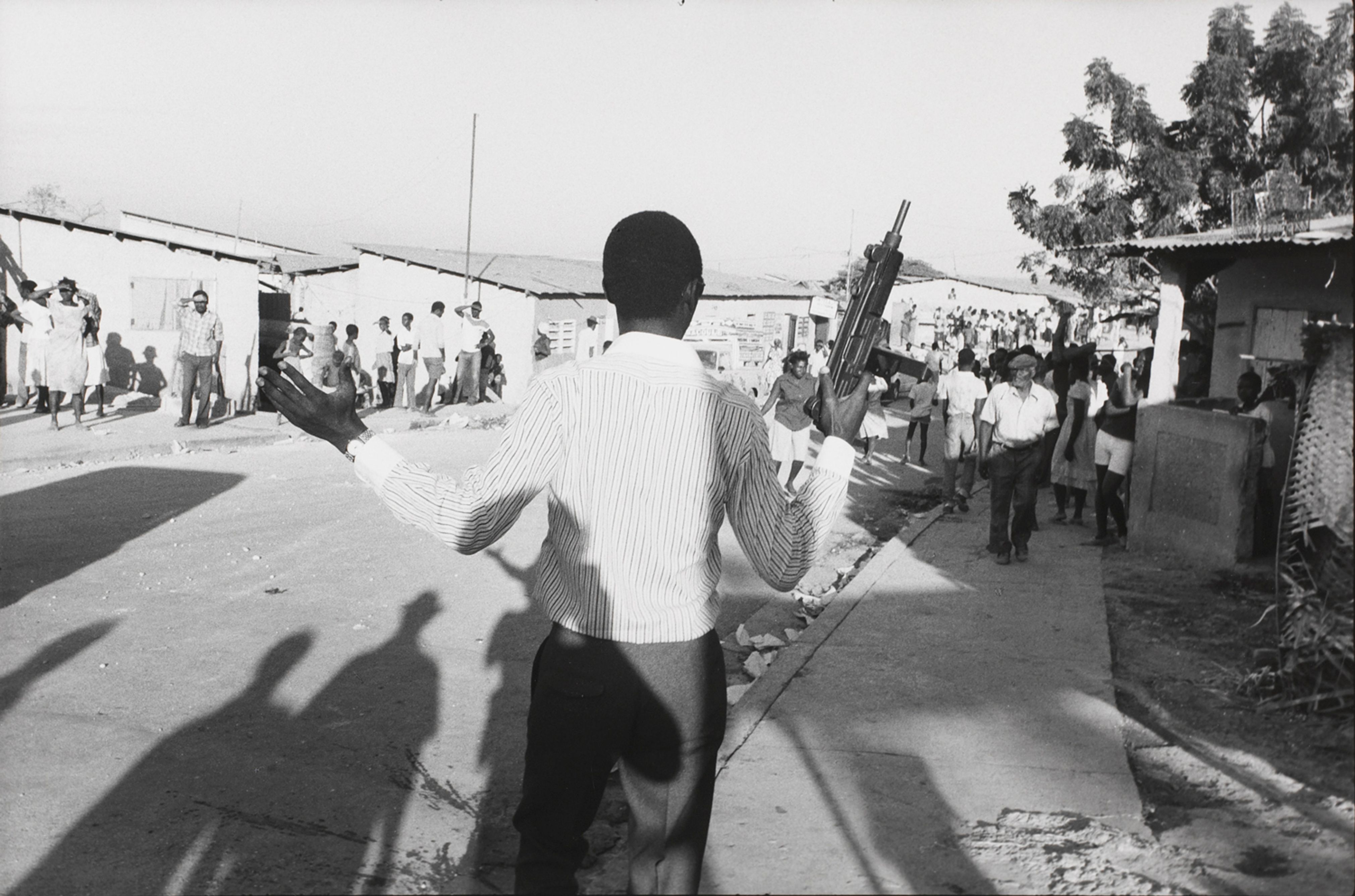 An out-of-uniform Tonton Macoute with an Uzi machine gun harasses a crowd