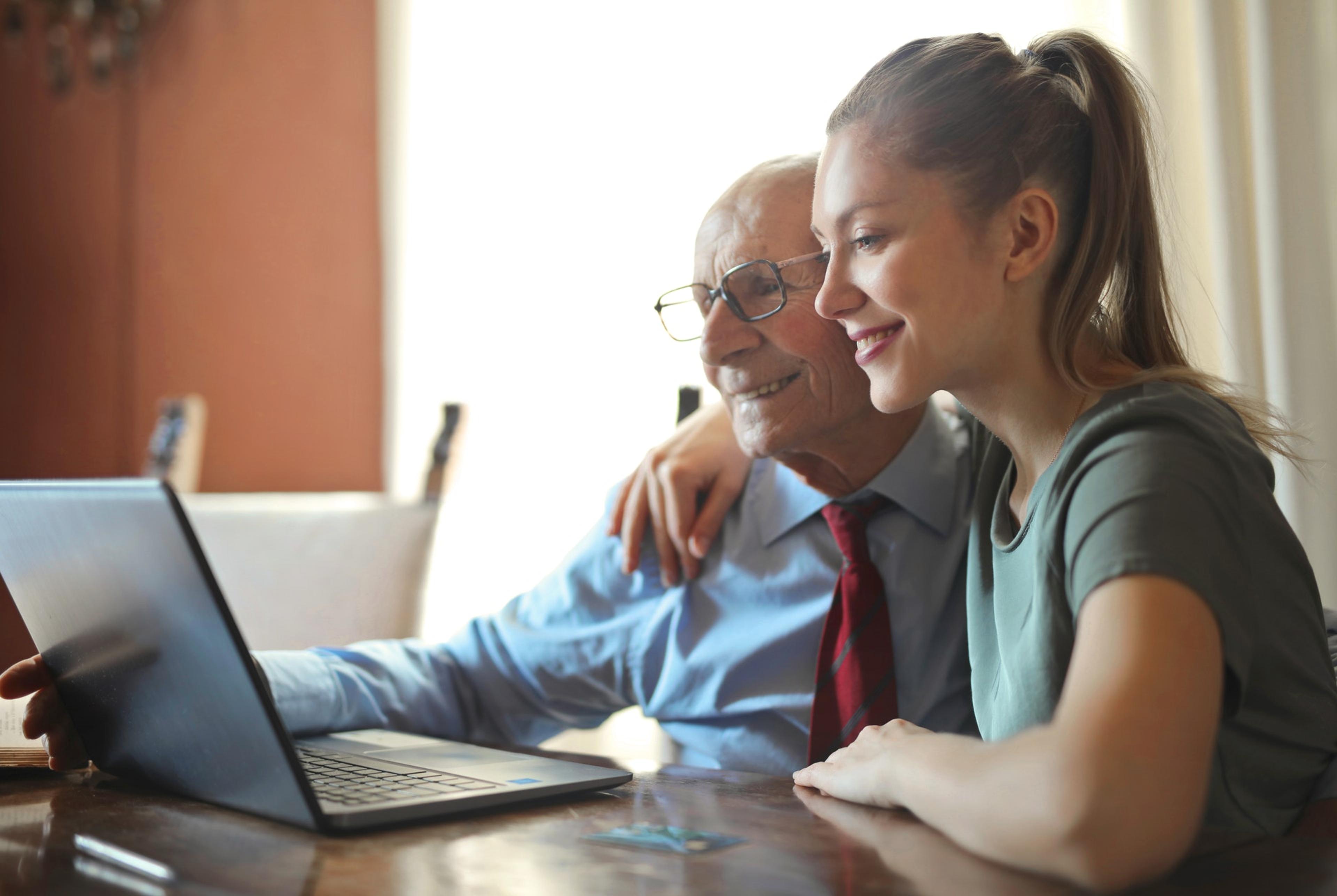 A young woman sits with an older man looking at a laptop, both smiling