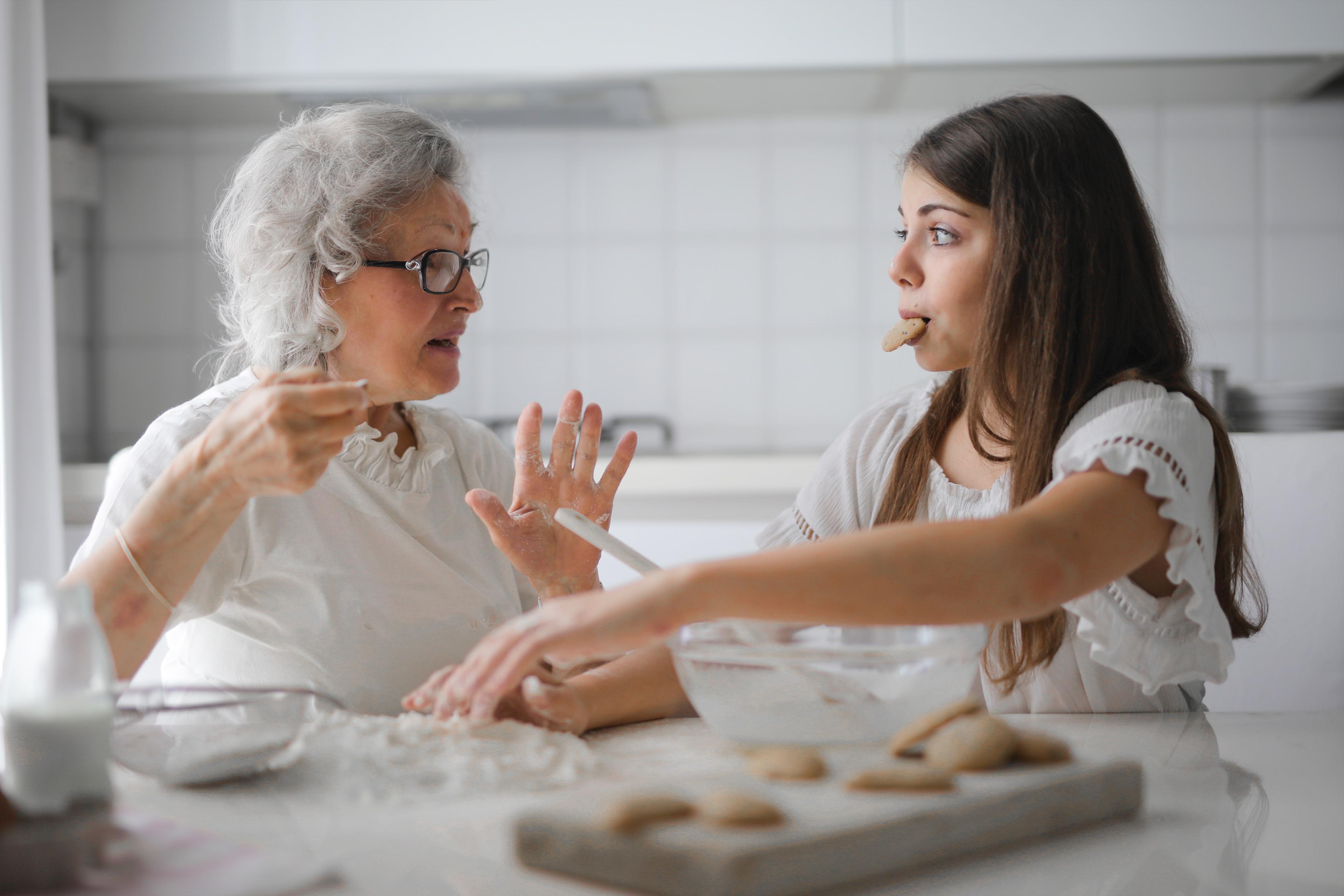 An older woman and a younger woman, both in white, sit at a table eating cookies