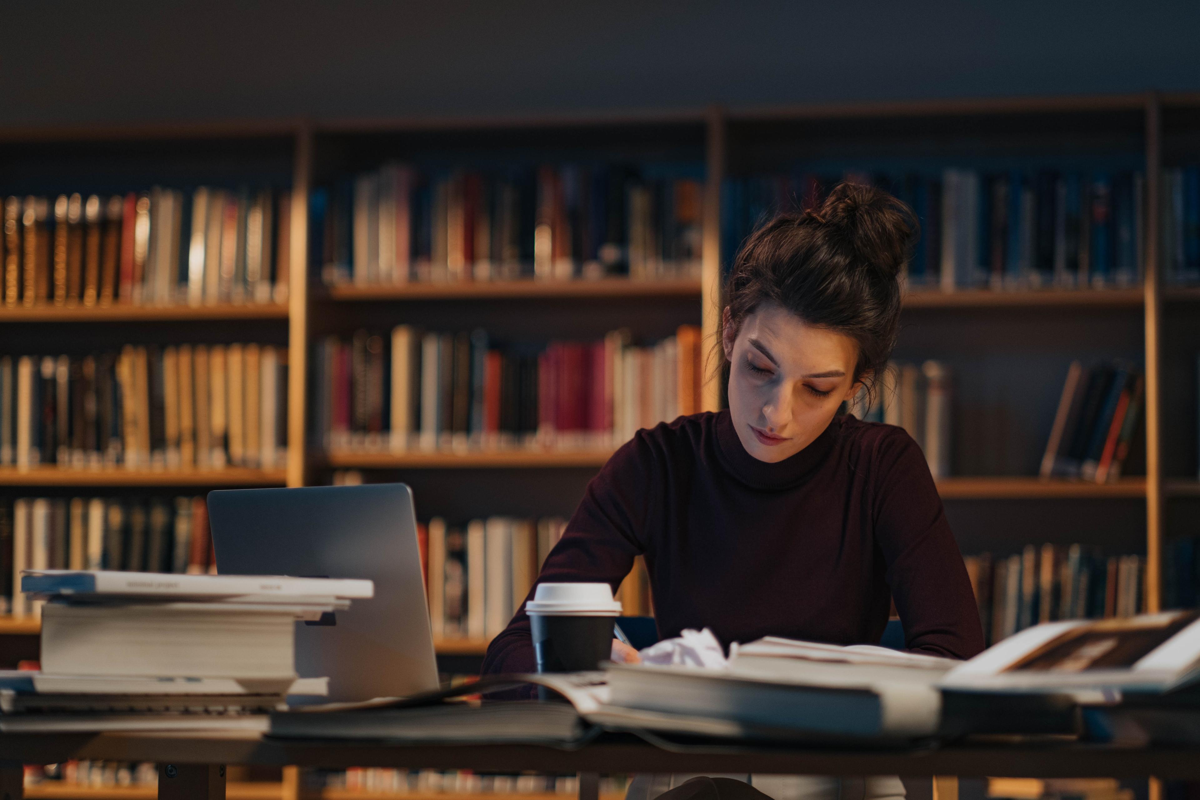A woman in a black turtleneck in front of an open laptop reads a book in a library