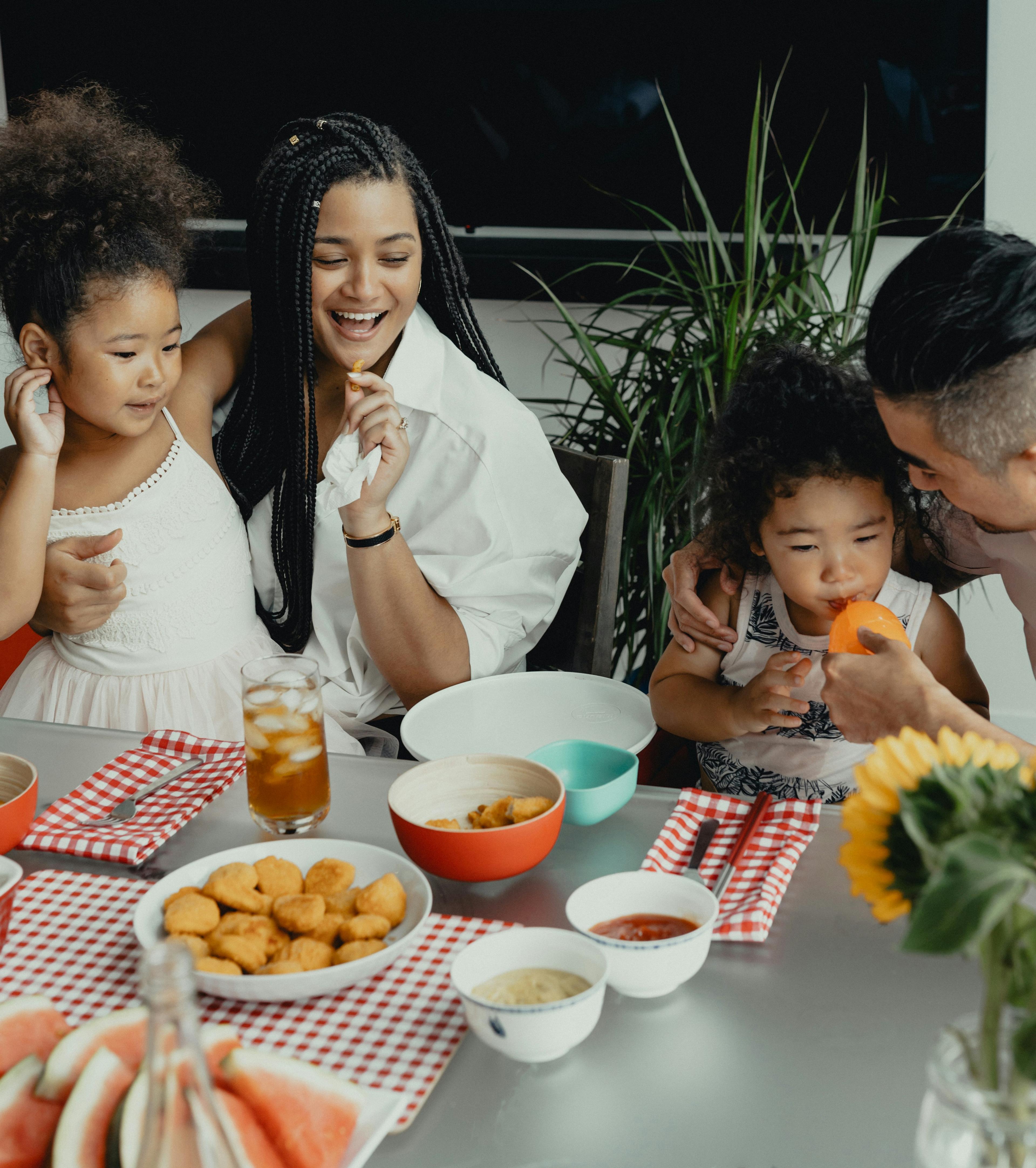 A family with two young daughters sits at a table eating a meal of chicken nuggets
