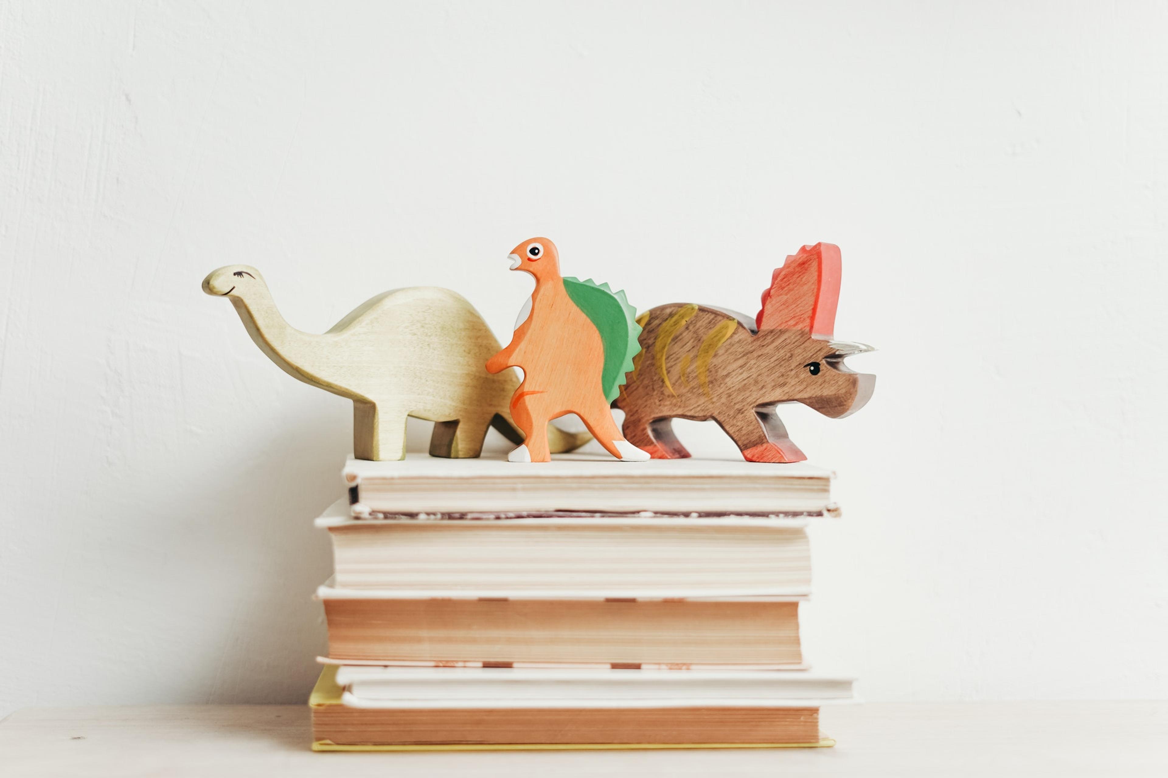 Three wooden dinosaurs atop a stack of books against a white background