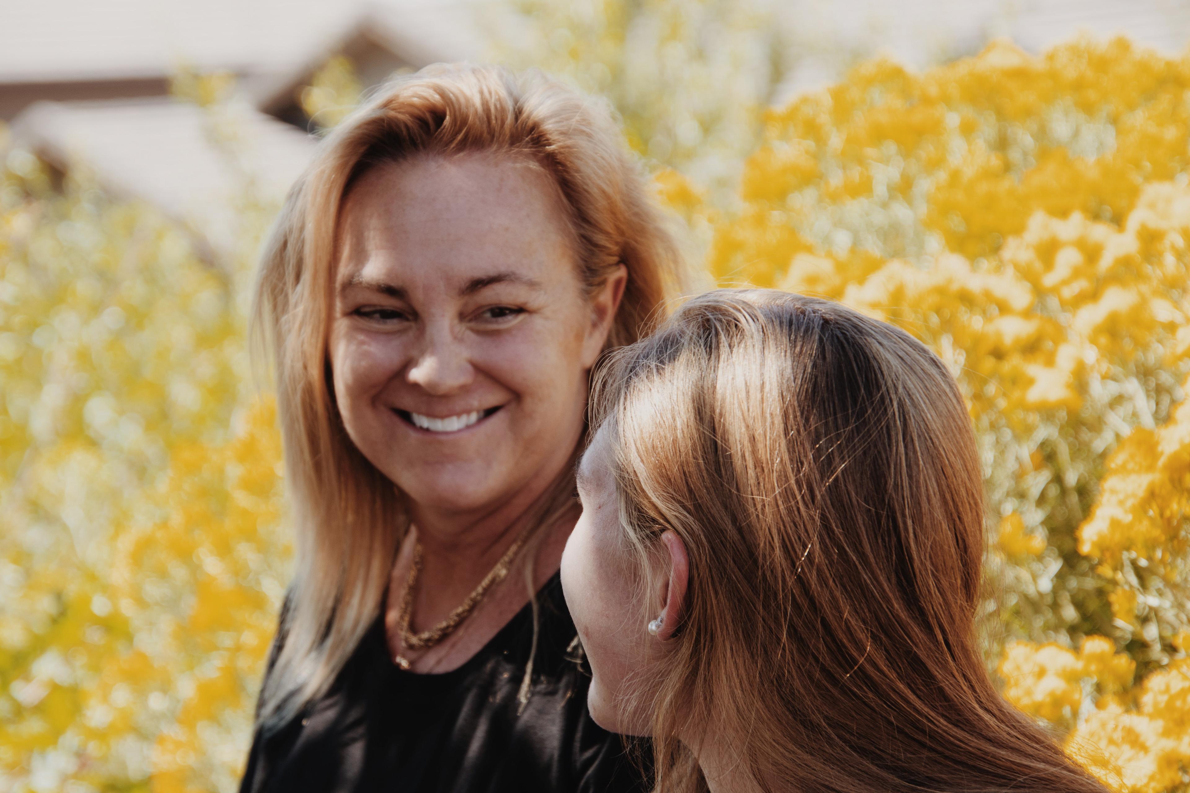 A mother smiles at her adolescent daughter in front of a yellow bush