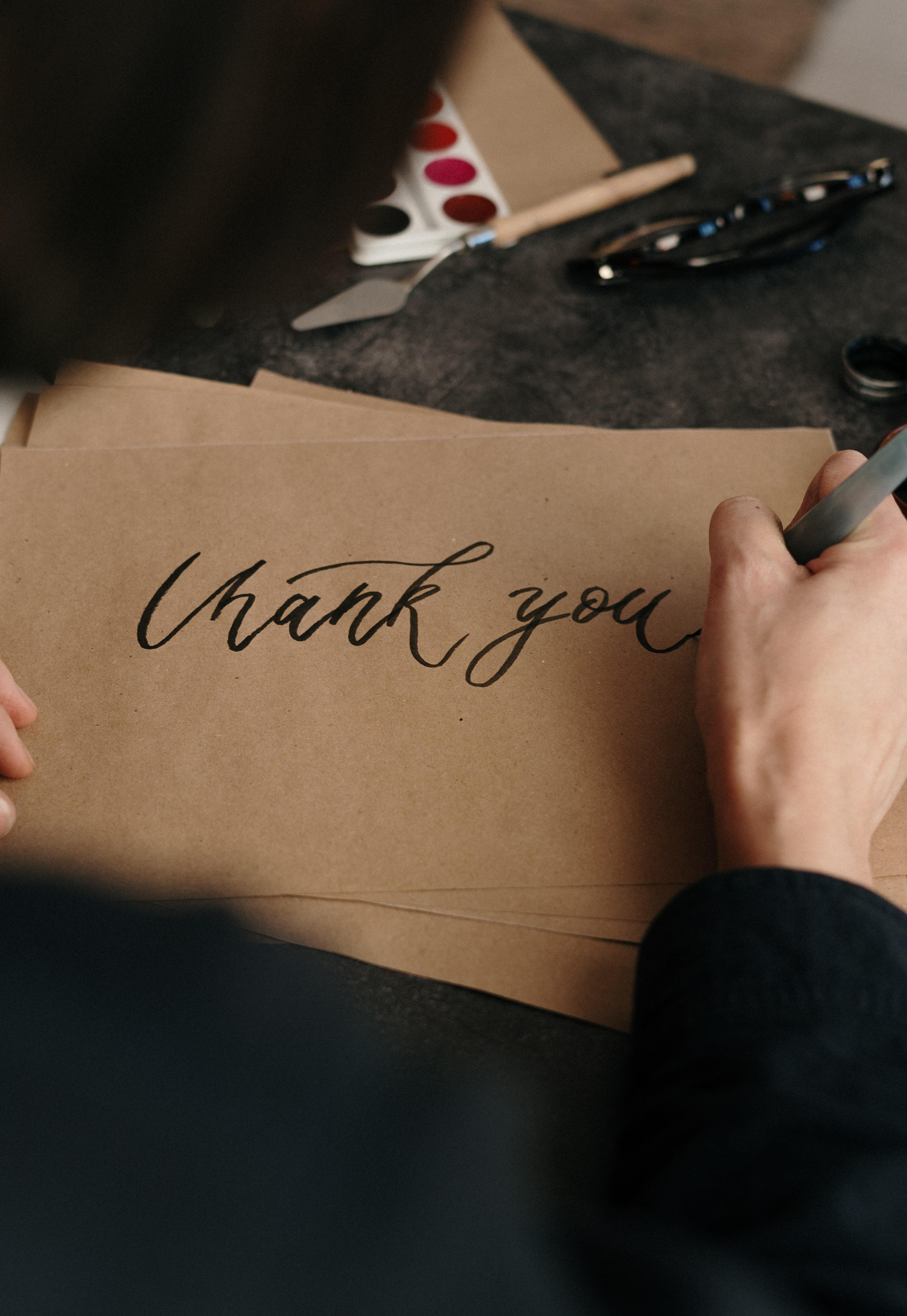 A hand with a pen writes "thank you" on a brown piece of paper