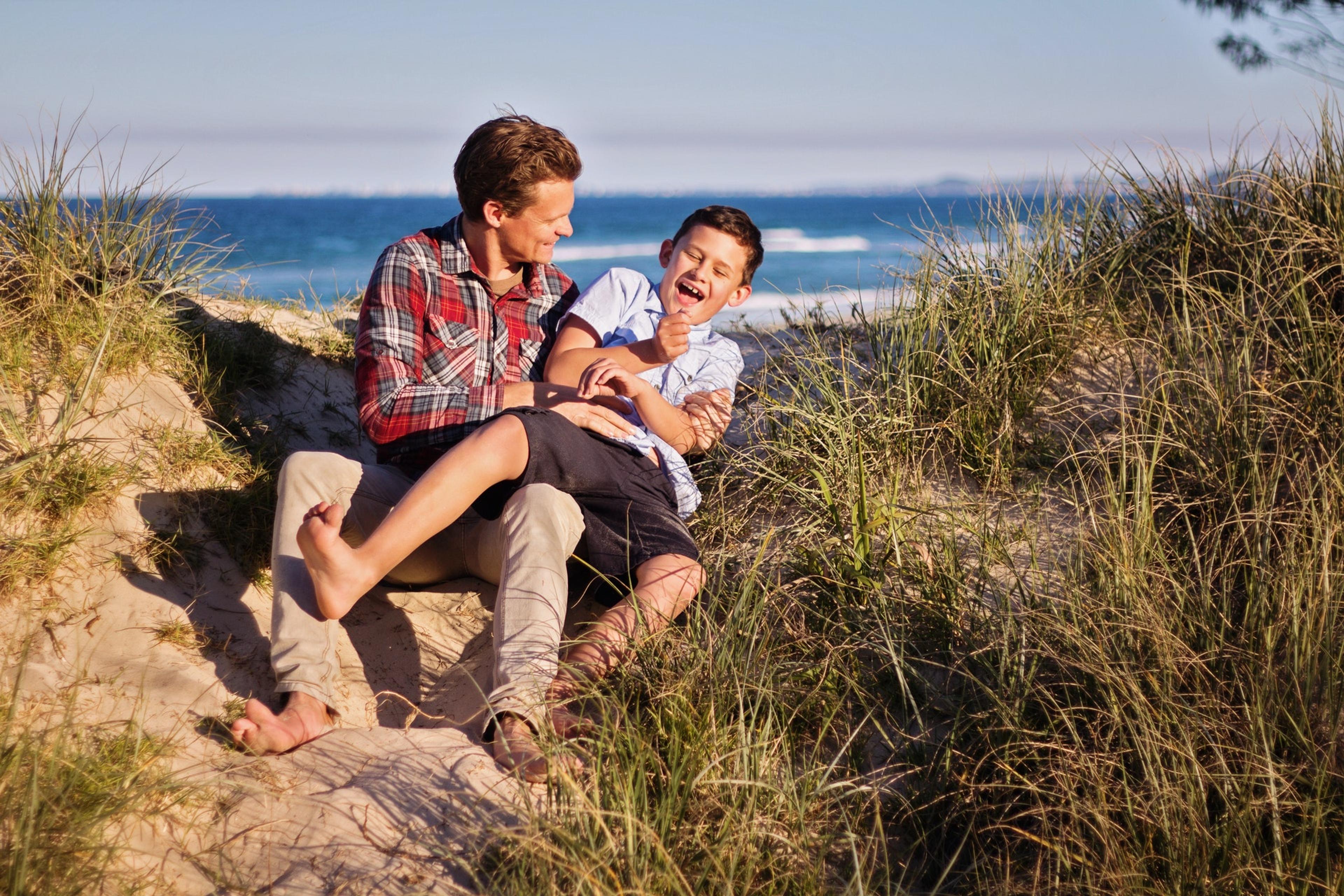 A man and a young boy laugh while sitting together on a sand dune