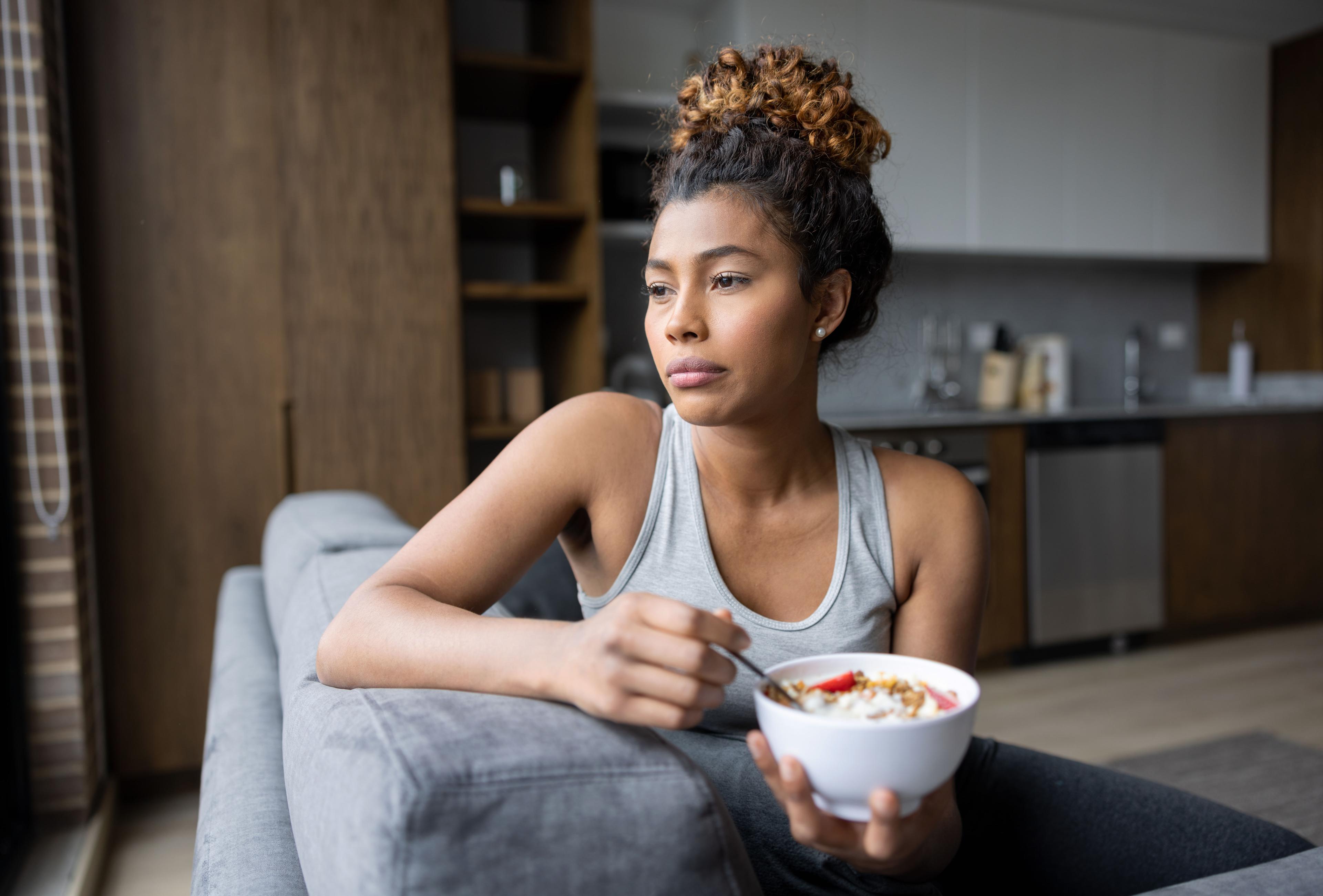 A woman in a tank top sits on a couch, holding a bowl of cereal, looking sadly into the distance