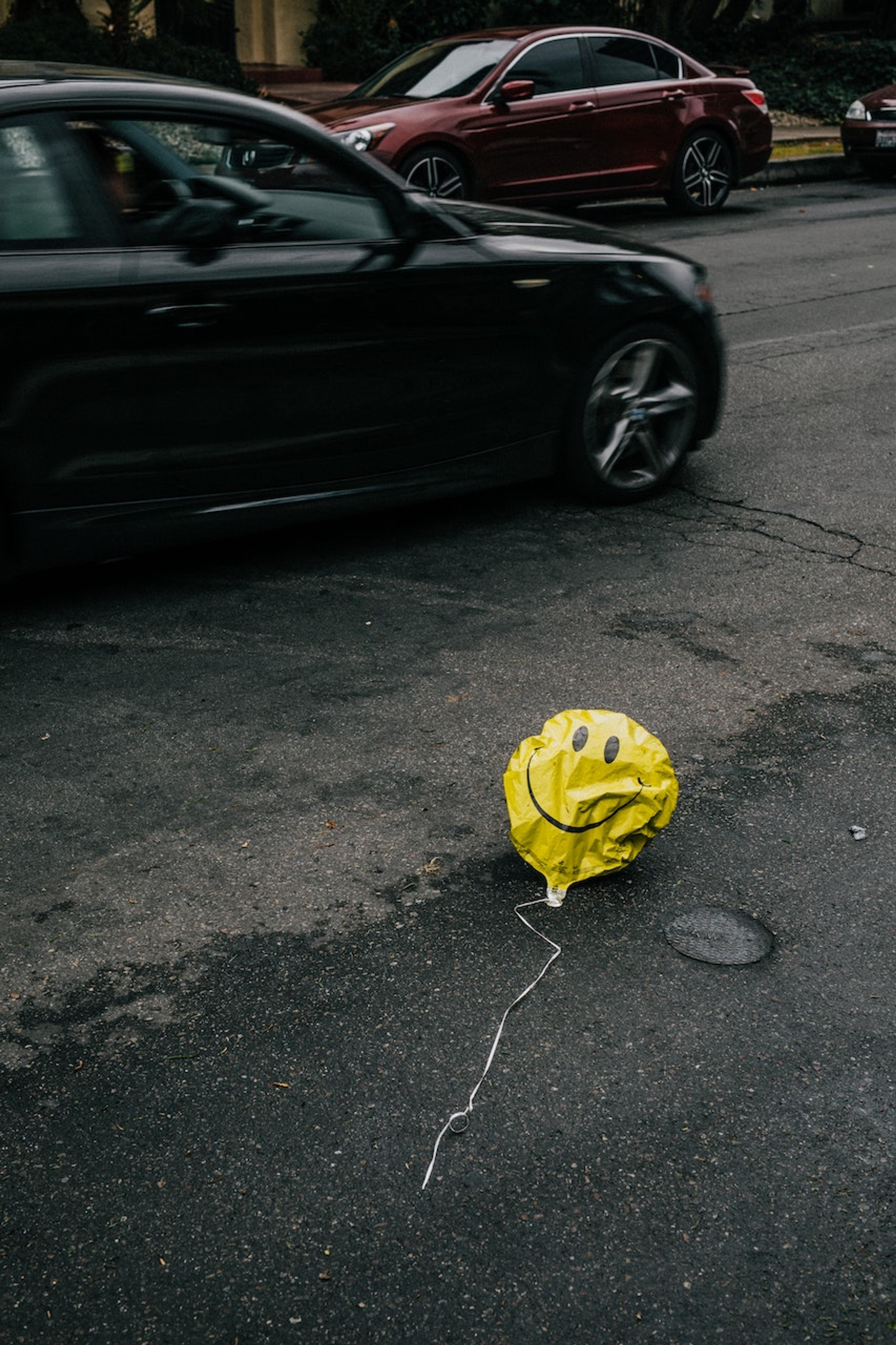 A deflated yellow smiley-face balloon blows along the pavement while cars drive by